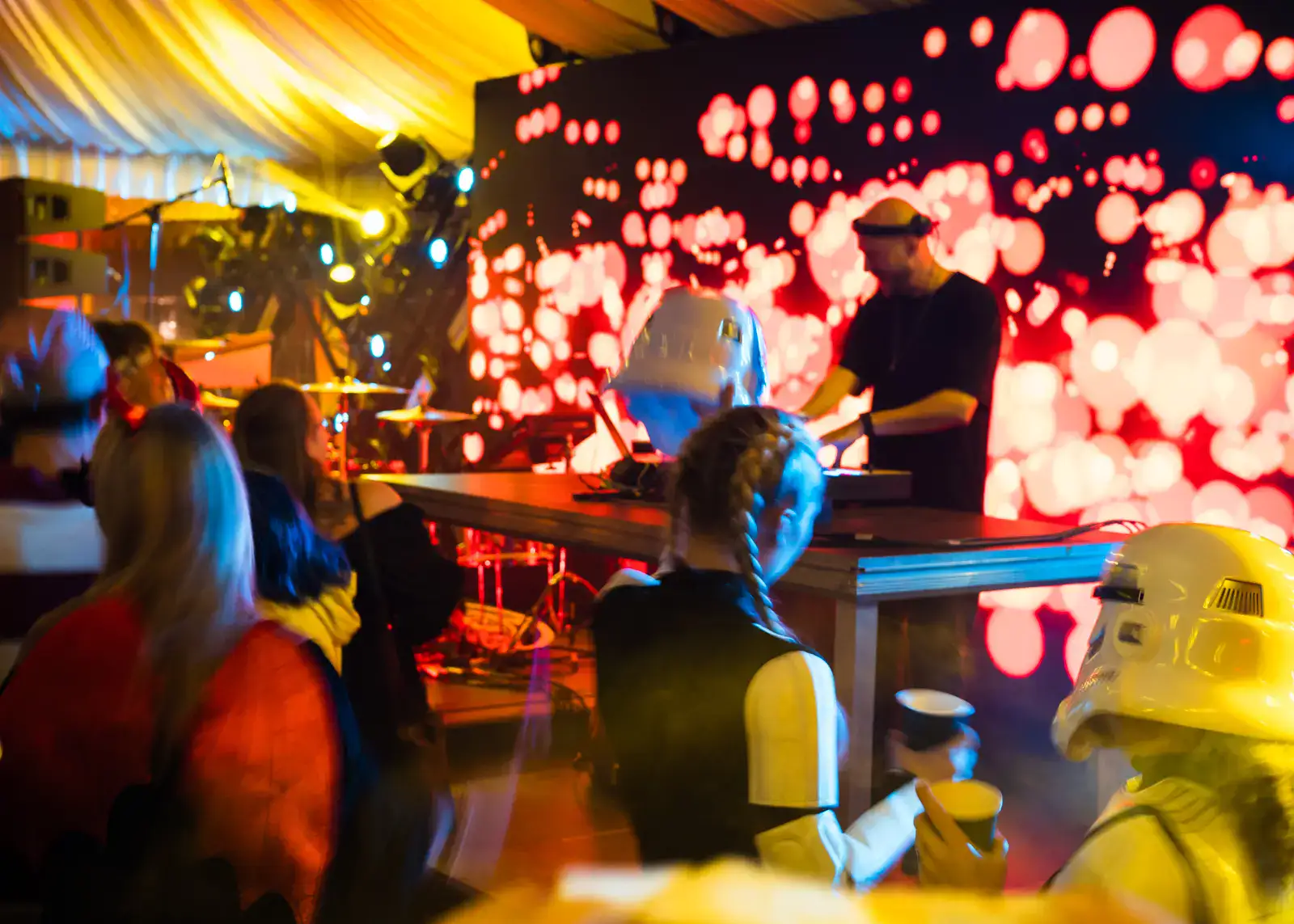 Dj in black clothes on a stage with red and white light dots in the background, people in costumes in front, yellow lights, at the Bran Castle Halloween party.