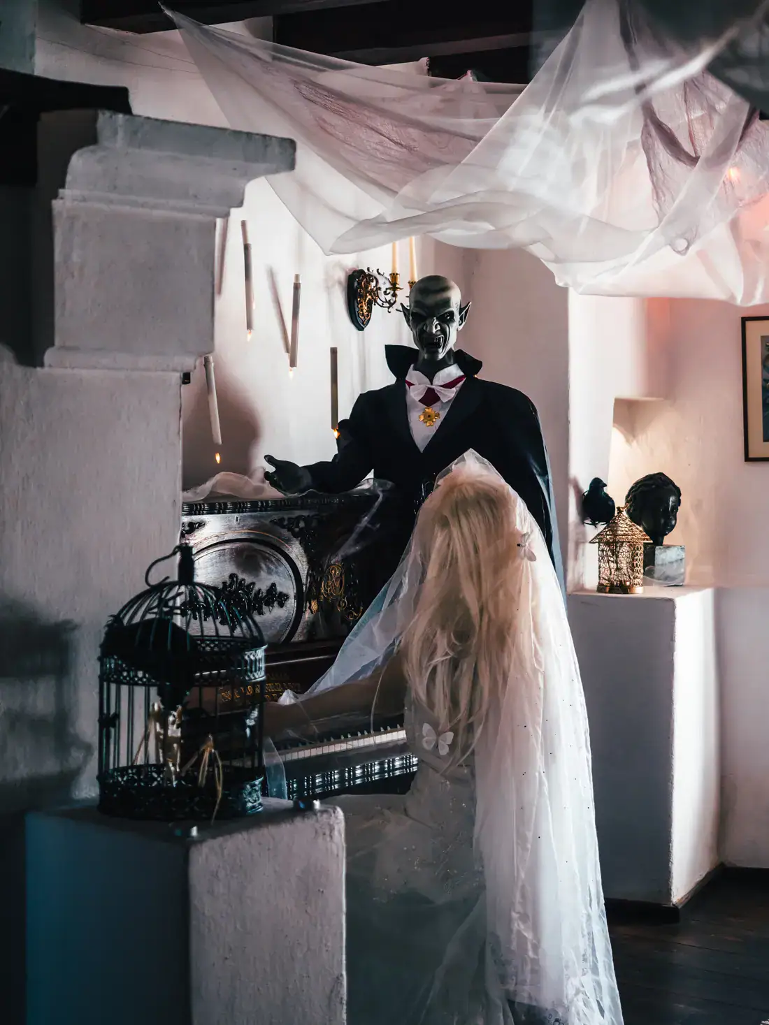Life-size Dracula figure in a black tux, next to a piano with a figure dressed in a bridal gown and veil, inside Bran Castle, decorated for Halloween.