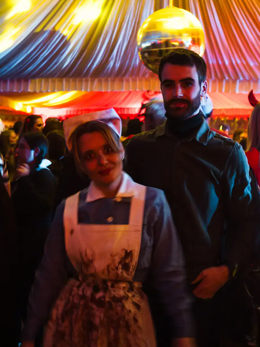 Woman in old nurse costume, with a grey dress and white apron with blood on it, taller man with dark hair and beard, wearing dark clothes, inside the tent at Bran Castle Halloween party.