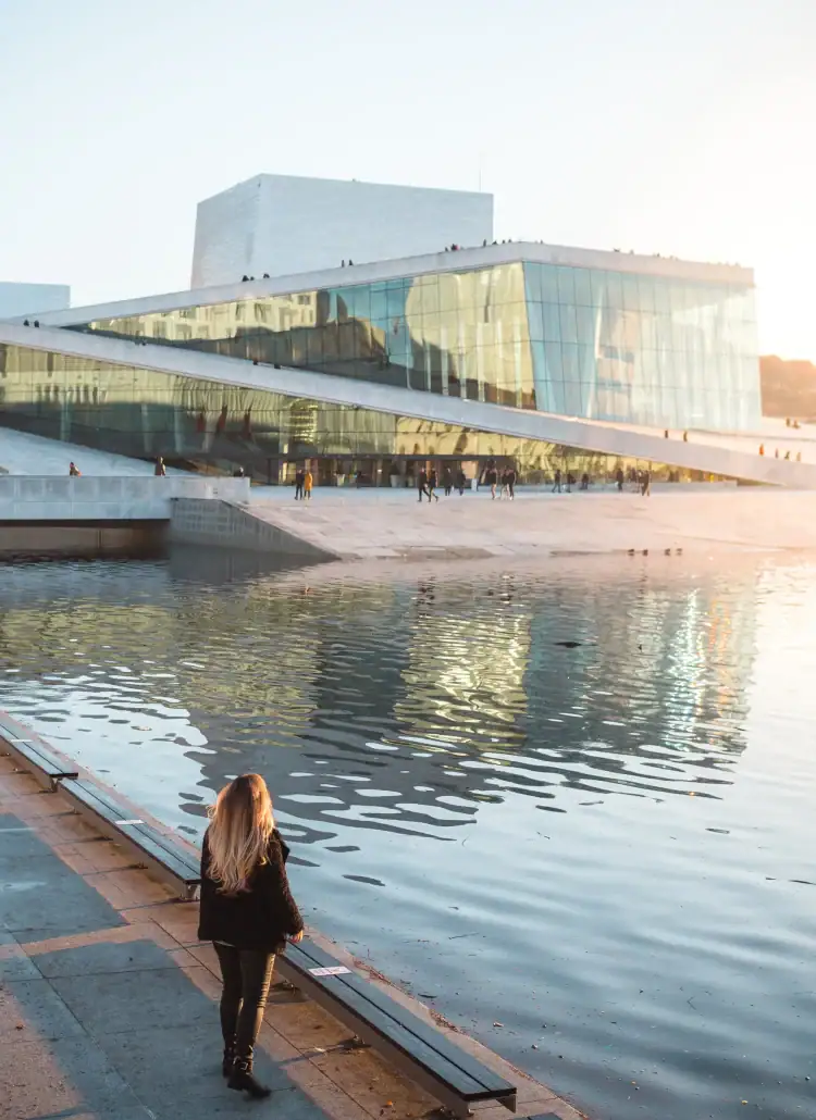 Woman with long, dark blonde hair, wearing a black jacket and pants, standing by the waters edge with the modern, white Oslo Opera House in the background at sunset. Where to stay in Oslo.