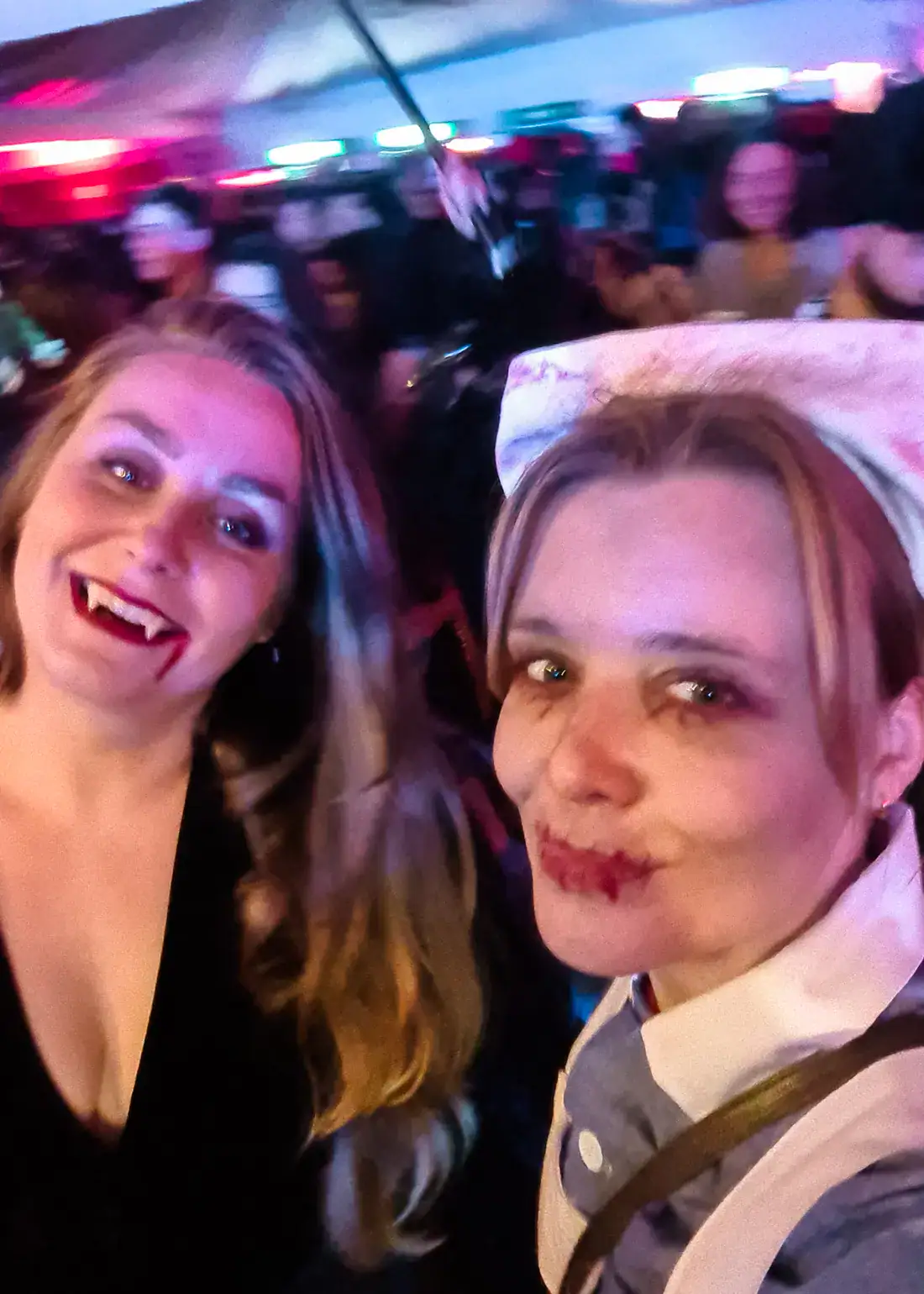 Woman with long hair, smiling with vampire teeth and woman as a dead nurse in old uniform smirking to the camera, at the Bran Castle Halloween Party.