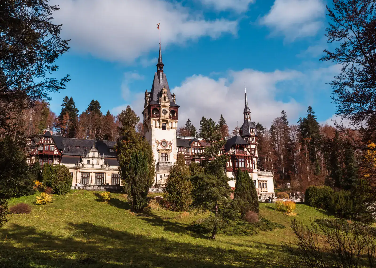 View across a green field of Peles Castle, white and dark brown timber, with one large tower in the middle, smaller turret on the right, seen on a lightly cloudy day traveling from Brasov to Bucharest with Daytrip.