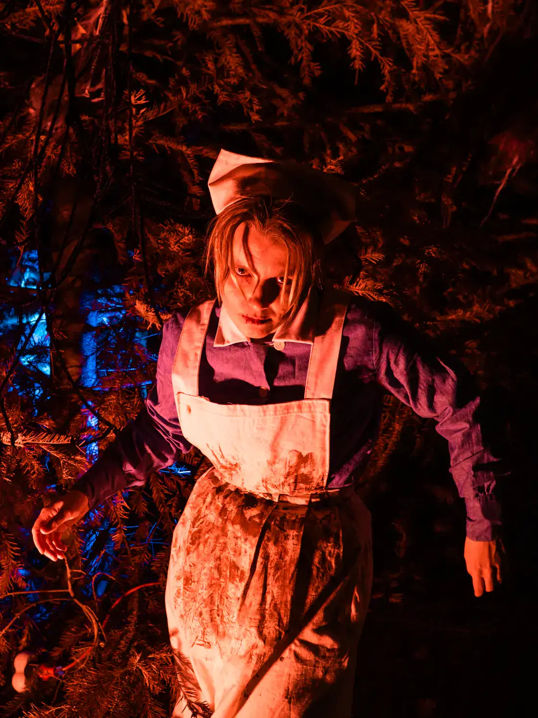 Woman wearing an old nurse uniform, grey dress with a white apron and hat, covered in blood, holding arms in a weird position, lit up by red light at the Halloween party at Bran Castle.