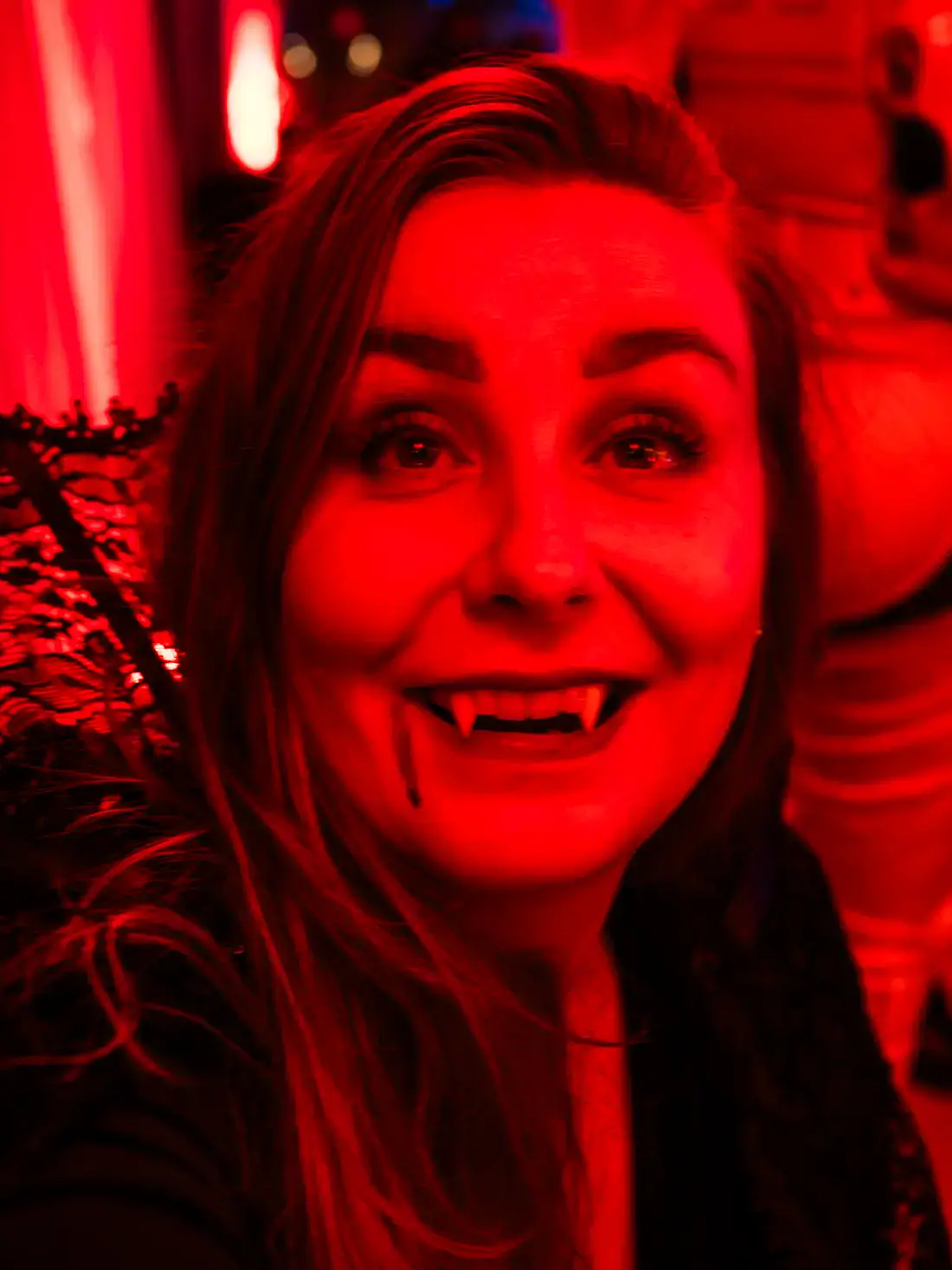 Close up of woman with long hair dressed as a vampire, smiling with fangs, looking up over the camera, in red light at Bran Castle Halloween party.