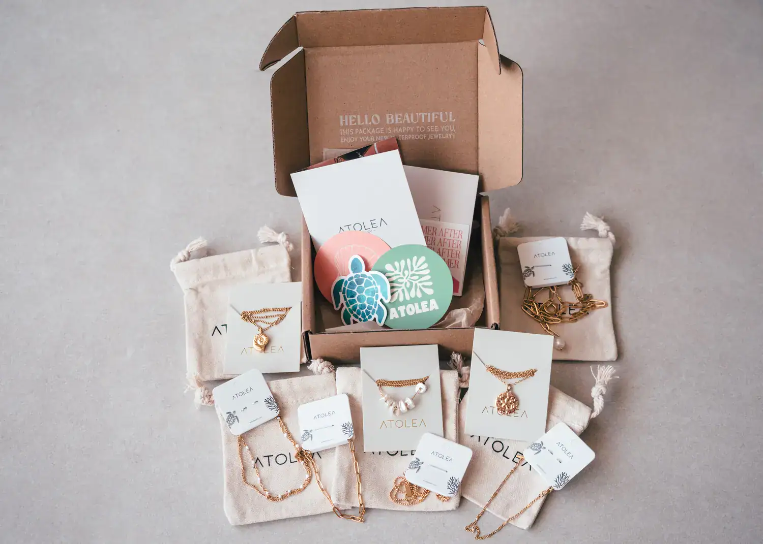 Display of a small brown cardboard box, surrounded by small beige pouches with gold necklaces and bracelets on top, beachy stickers from Atolea, my favorite travel jewelry.