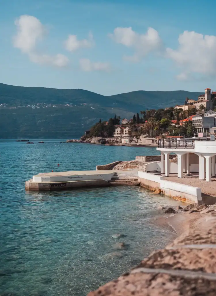 Looking from the waterfront promenade in Herceg Novi on a cement quay in shallow turquoise water, with Herceg Novi Old Town in the background on a sunny day.