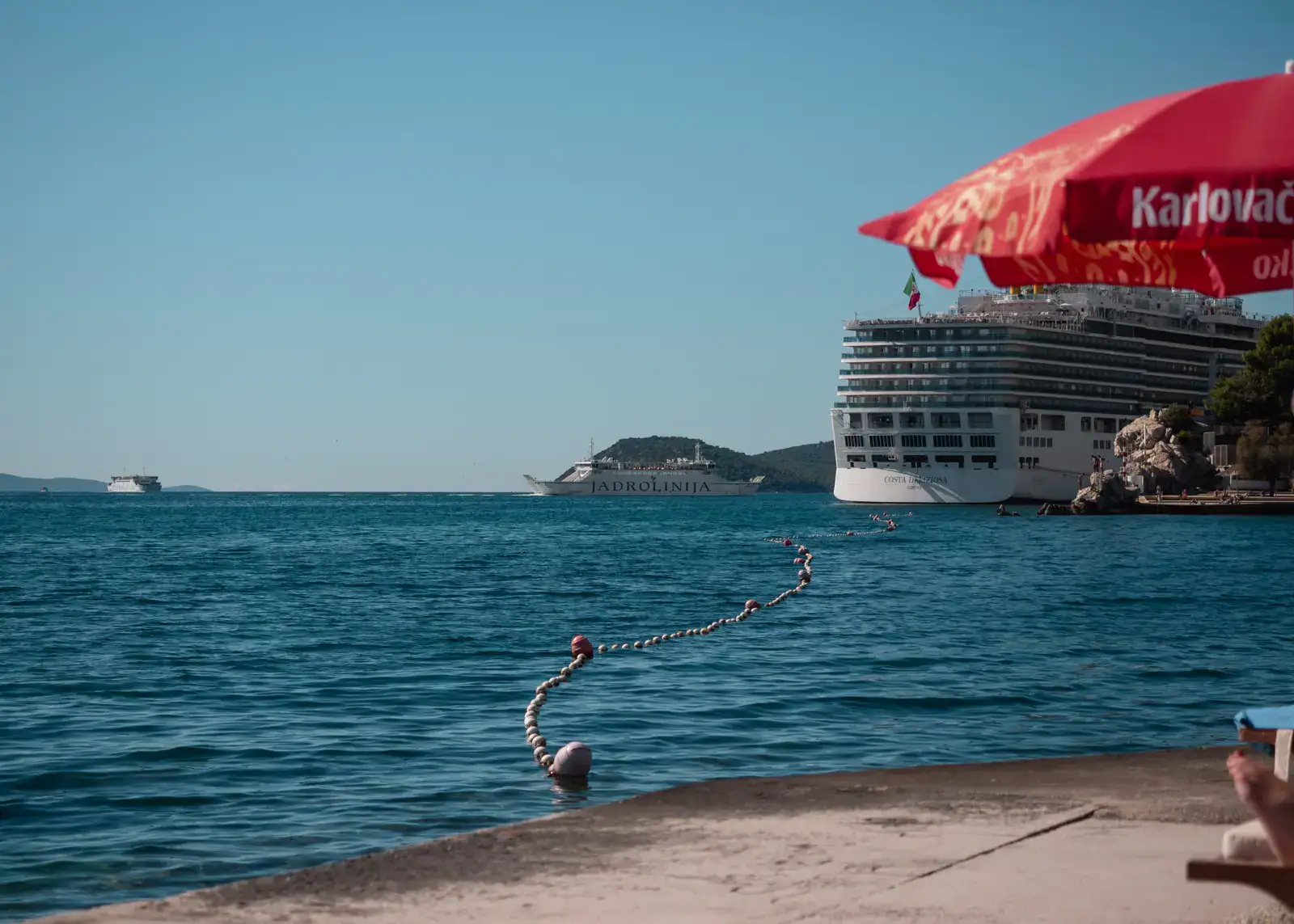 View from a concrete quay with a red parasol, of the end a large, white cruise shit docked at Split port, a smaller ferry going out, blue water and blue skies.