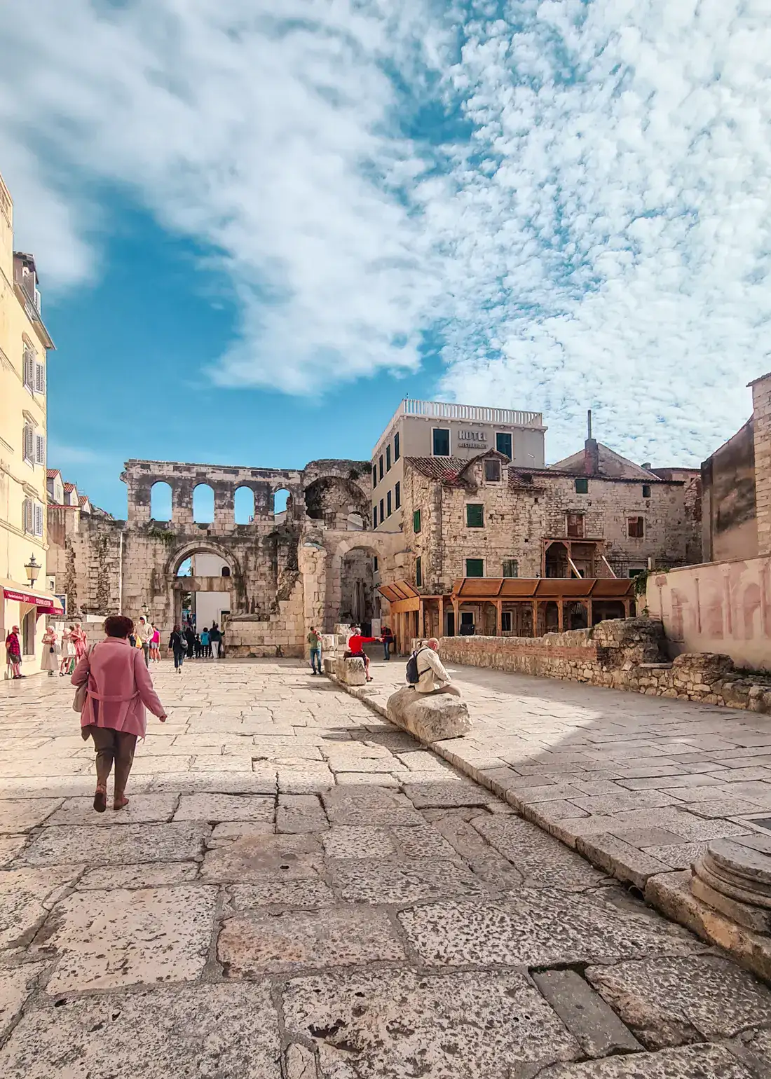 Walking behind a woman with a pink jacket inside Split Old Town surrounded by Roman ruins on a sunny day with some clouds. Reasons to visit Split.