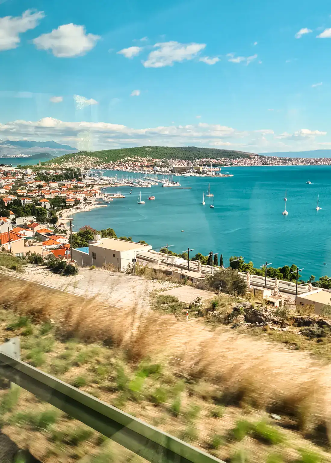 Turquoise bay with some sail boats, green island in the background, small houses with orange roofs, seen from a bus on my way to Split.