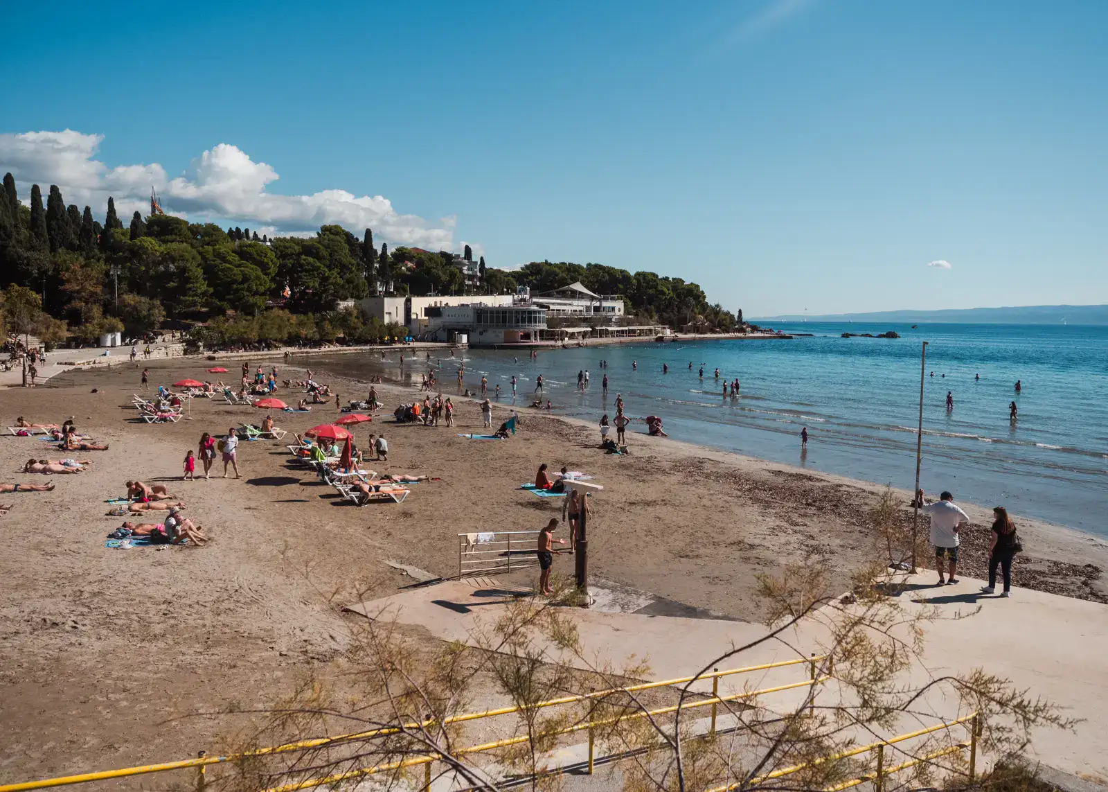 Looking out over Plaža Bačvice, the city beach in Split. Some people on sunbeams with red parasols, light brown sand backed by greenery, shallow, blue water on a sunny day in Split with blue skies.