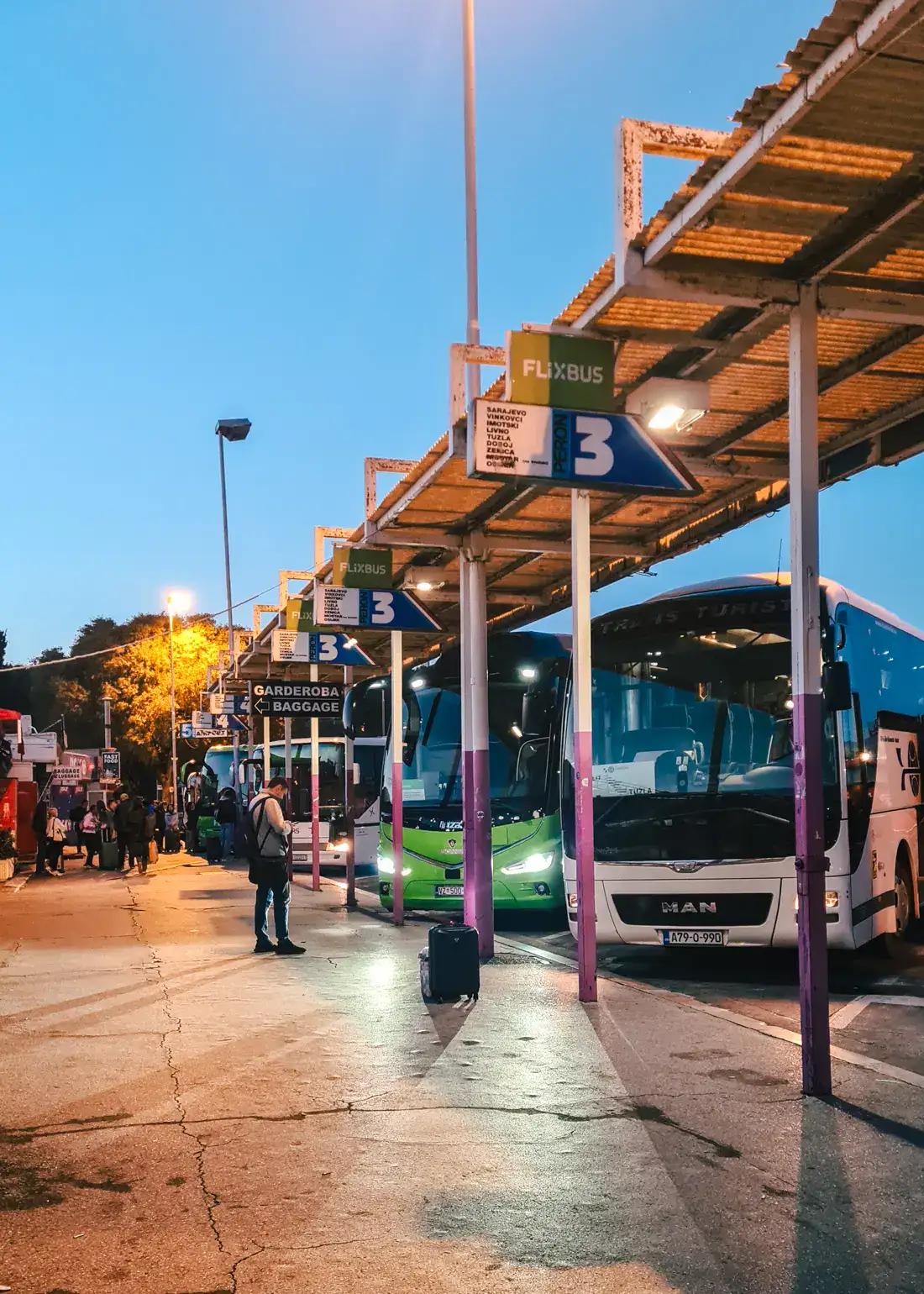 White and green buses lined up under a roof with white and pin pillars, street lights are on early in the morning at Split Bus Station.