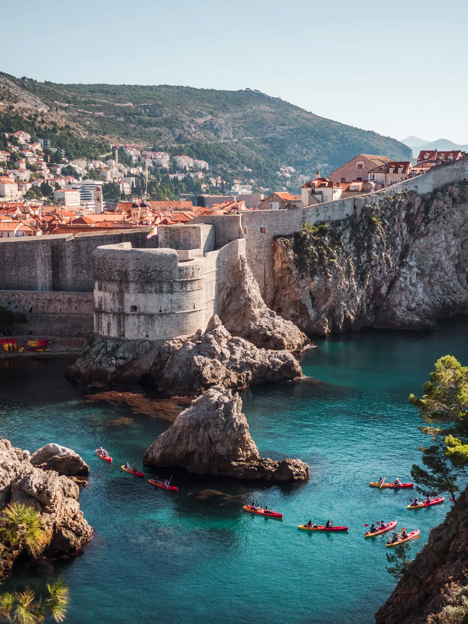View from Fort Lovrijenac of Dubrovnik walled Old Town with orange rooftops, turquoise ocean below the wall and 9 red and yellow double kayaks on a sunny morning, one of the highlights on any Dubrovnik itinerary.