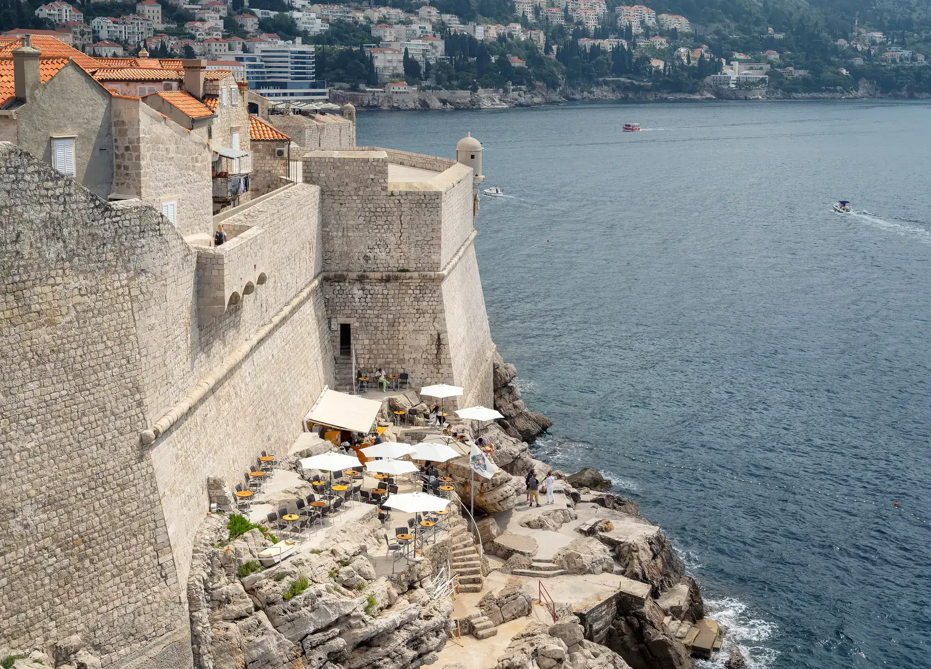 Buža Bar with white parasols built on different levels on a rocky outcrop on the outside of the tall Old Town walls on a sunny day in Dubrovnik.