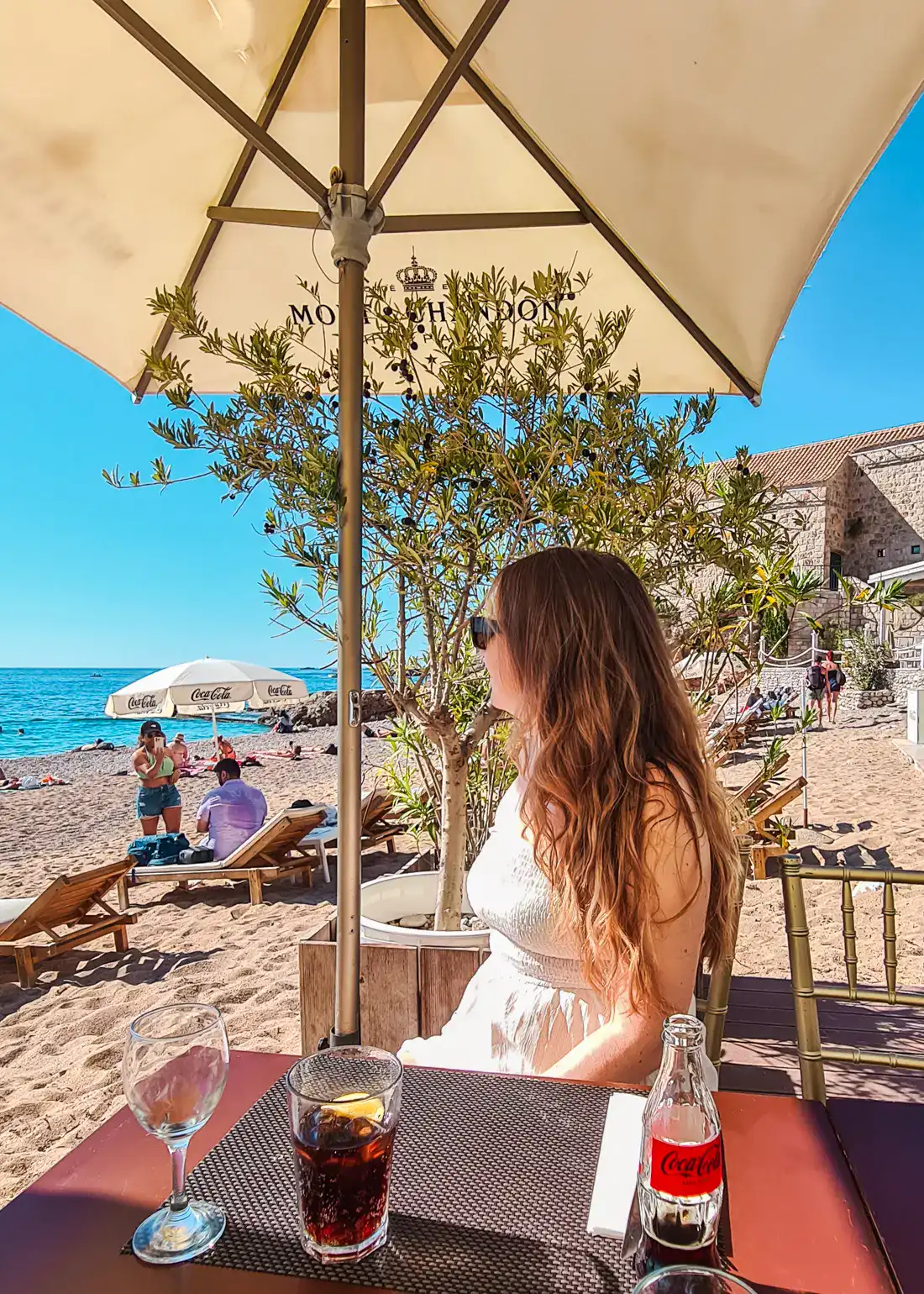 Woman with long brown hair, wearing a white dress, looking out over the Banje Beach and the ocean from a table at Banje Beach Club, under a beige parasol on a sunny day in Dubrovnik.