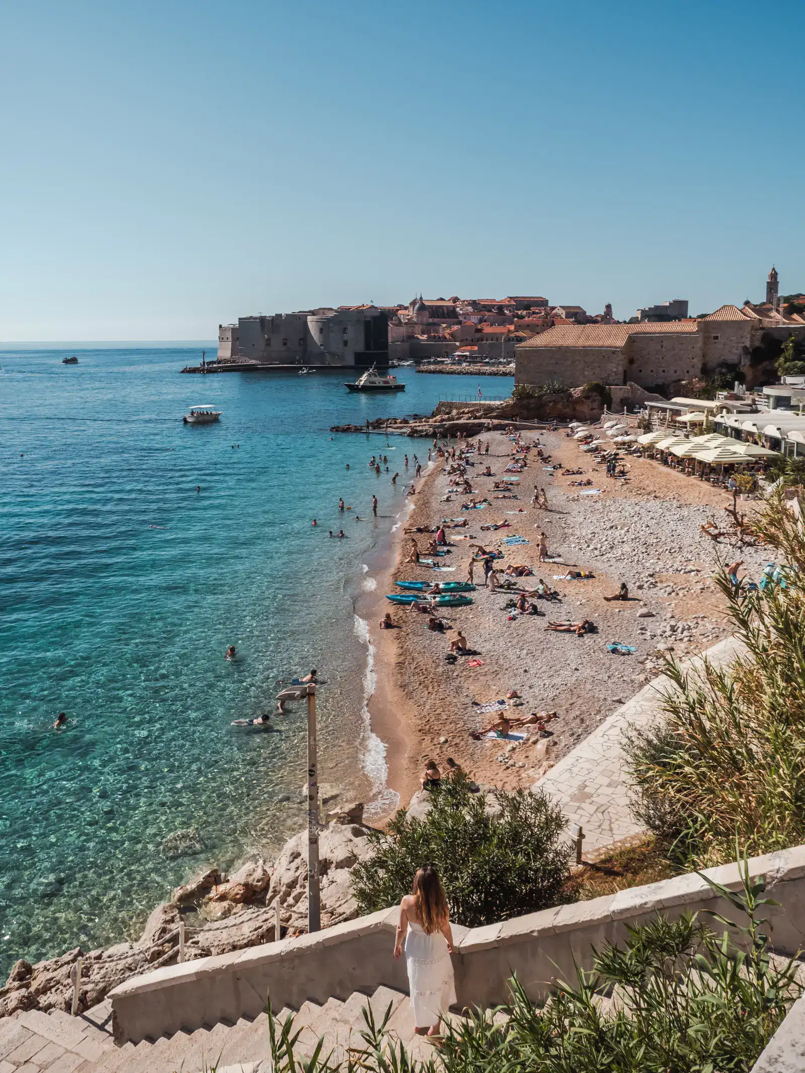 Woman in a white dress standing on stone stairs leading down to the ocean, looking over a beach with many people with Dubrovnik Old Town in the background and turquoise ocean in the left, one of the top things to do during a 3-day Dubrovnik itinerary.