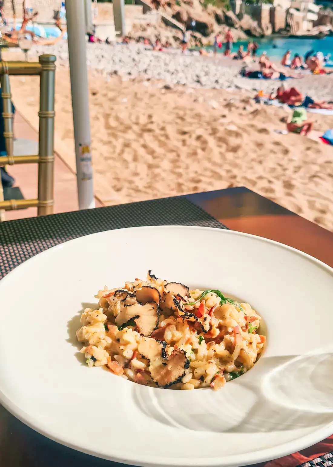 Round, white plate with truffle risotto on a wooden table on Banje Beach, a must during any Dubrovnik itinerary.