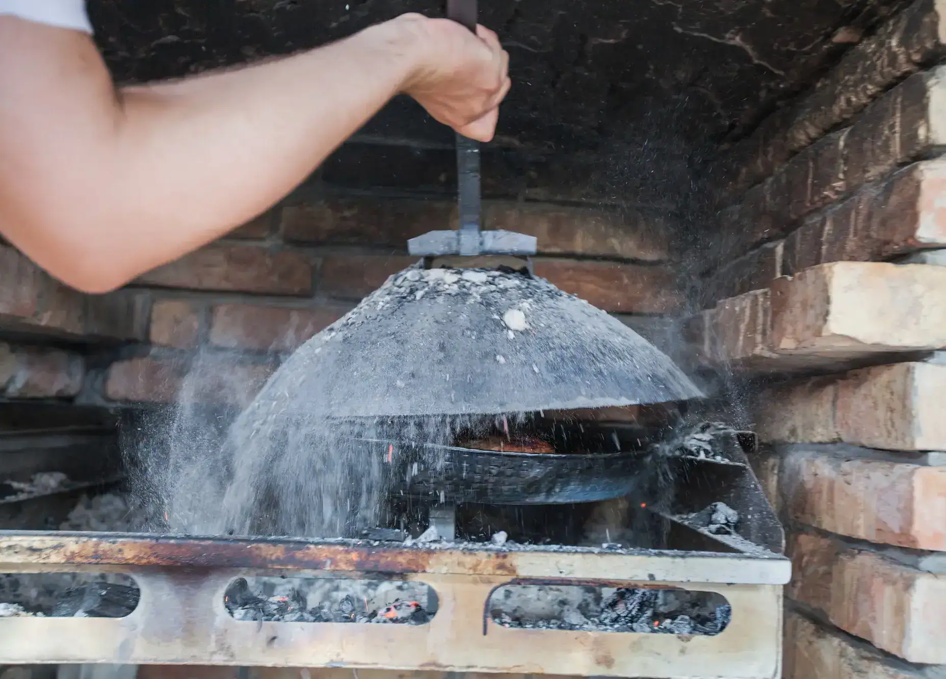 Hand holding a iron domed lid covered in ash inside a brick fire place, making Peka for dinner, a must-try during your Dubrovnik itinerary.