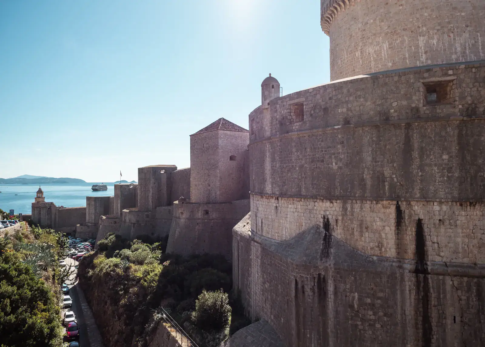 Looking on a rounded part of the grey Dubrovnik Old Town walls from the outside, with towers stretching to the ocean with a cruise ship in the distance on a sunny morning during a perfect Dubrovnik itinerary for first timers.