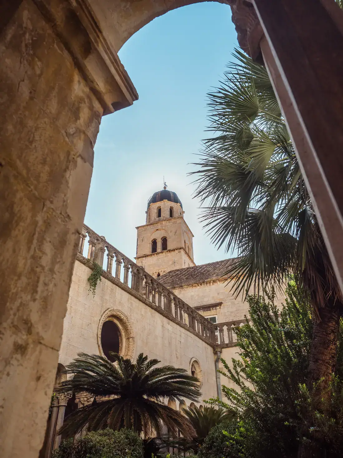 Looking through a stone arch up at a domed tower inside the Franciscan Church and Monastery with palm trees and greenery in the courtyard, on a sunny day with blue skies, during the perfect 3-day Dubrovnik itinerary for first timers.