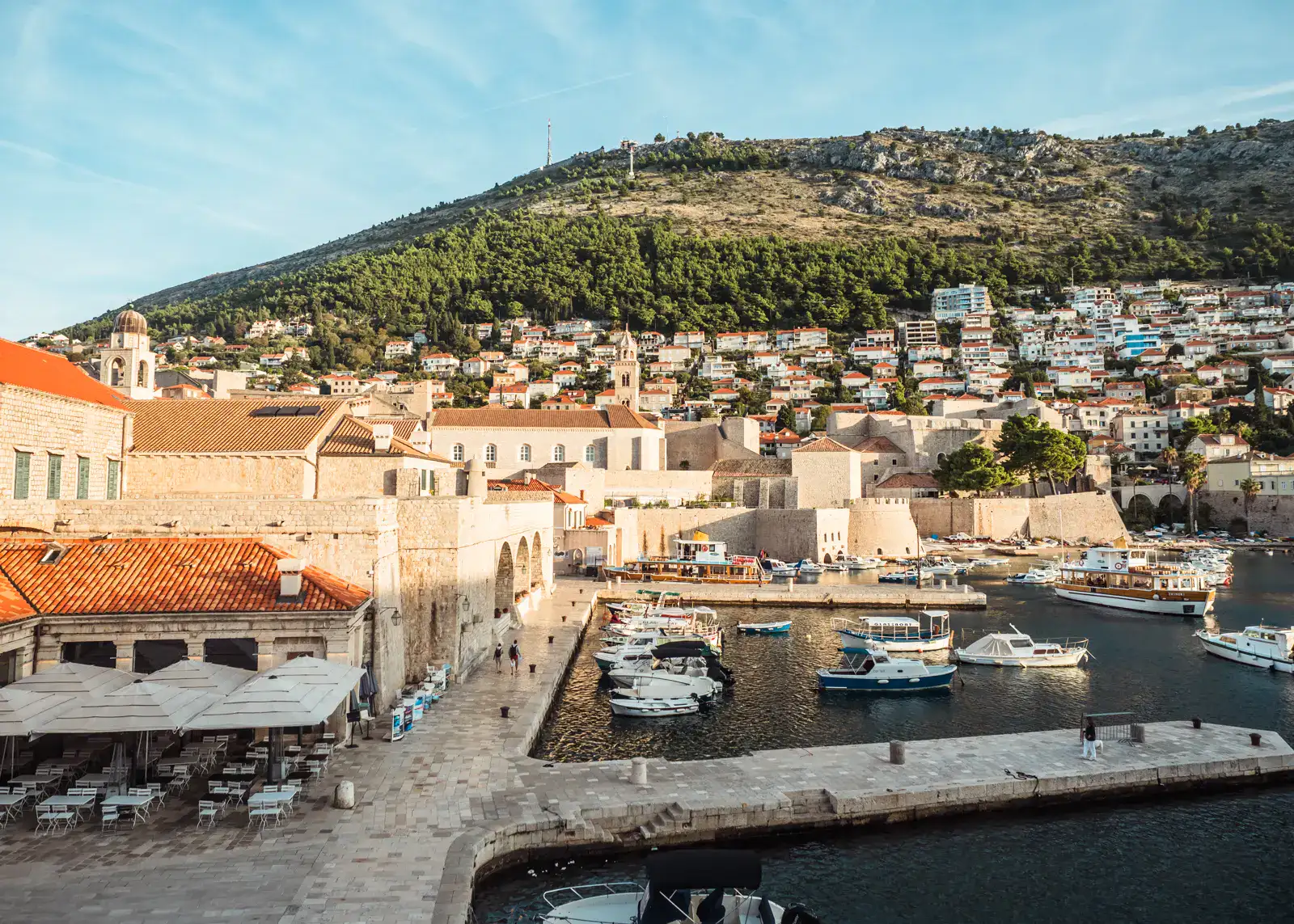 View from the Dubrovnik Old Town walls of the Old Harbor with restaurants on the left and Mount Srd in the background on a sunny morning in Dubrovnik - The perfect itinerary for first timers.
