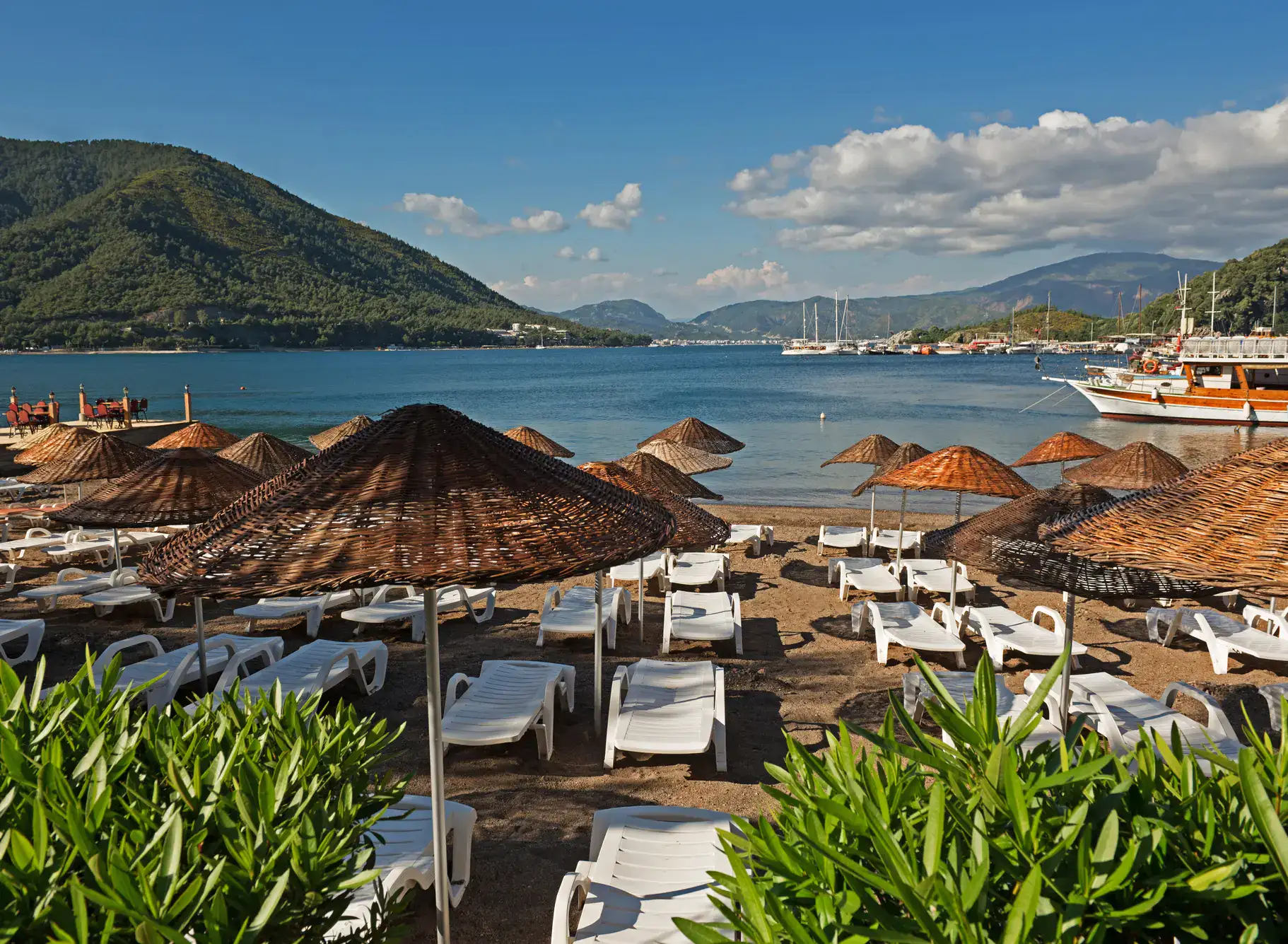 Beach with white sunbeams and brown, star umbrellas, green plants in the foreground, bay with calm, blue water and an island in the background in Marmaris, one of the cheapest beach destinations in T&uuml;rkiye.