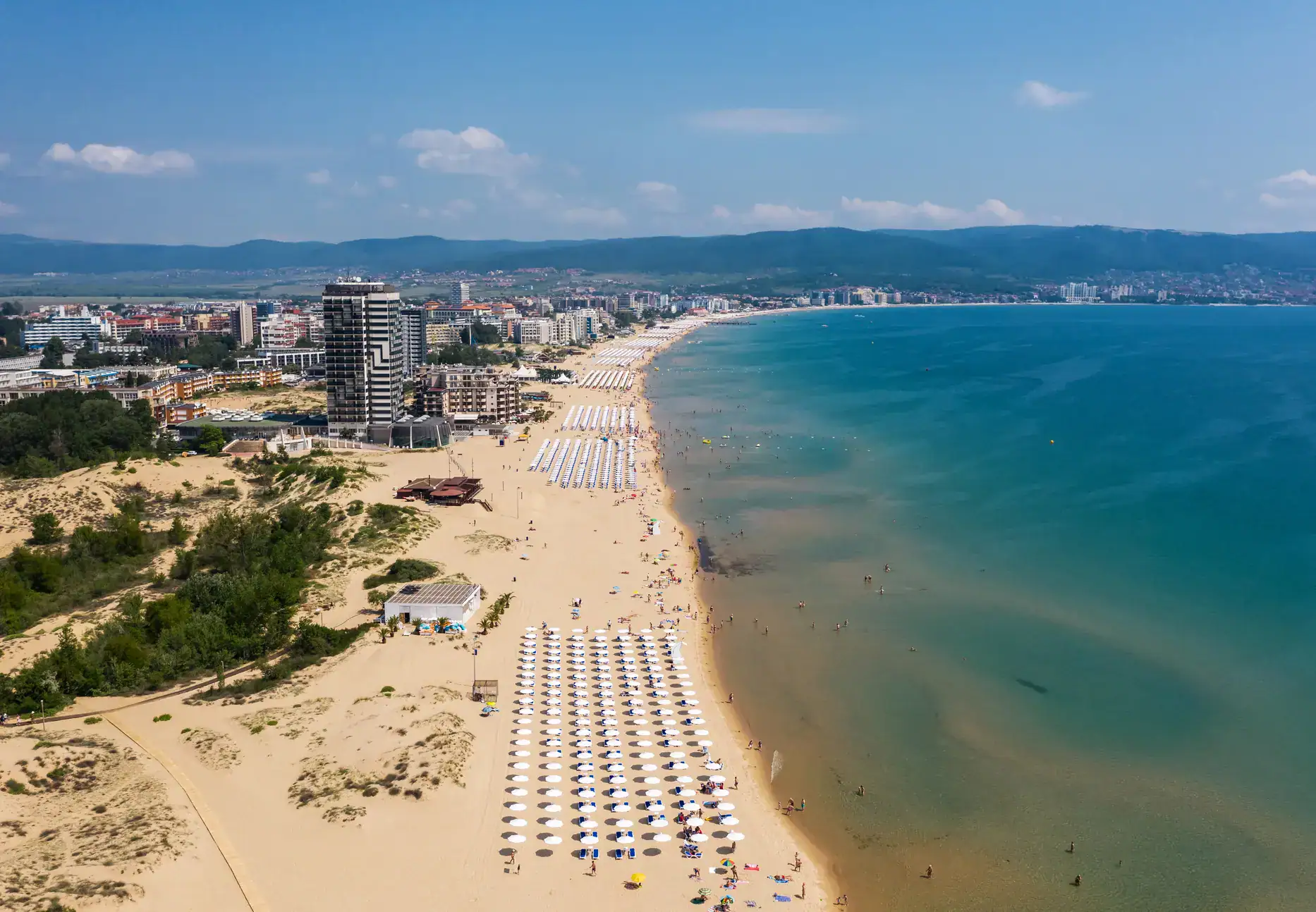 Drone view of Sunny Beach, a mile-long stretch of light sand with sections of sunbeams and white parasols, high-rise building along the beach on a sunny day. One of the cheapest beach destinations in Europe.