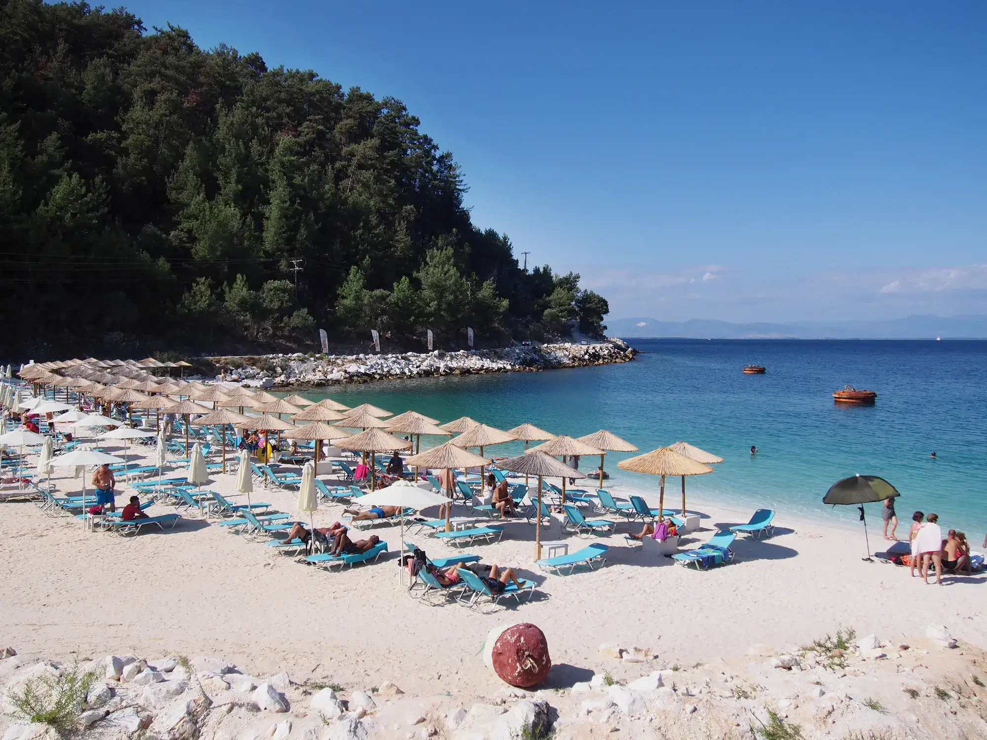 Marble Beach, a white, sandy bay with blue sunbeds and beige straw parasols, headland covered in green trees in the background on a sunny day with blue skies in Thassos, one of the cheapest beach destinations in Europe.