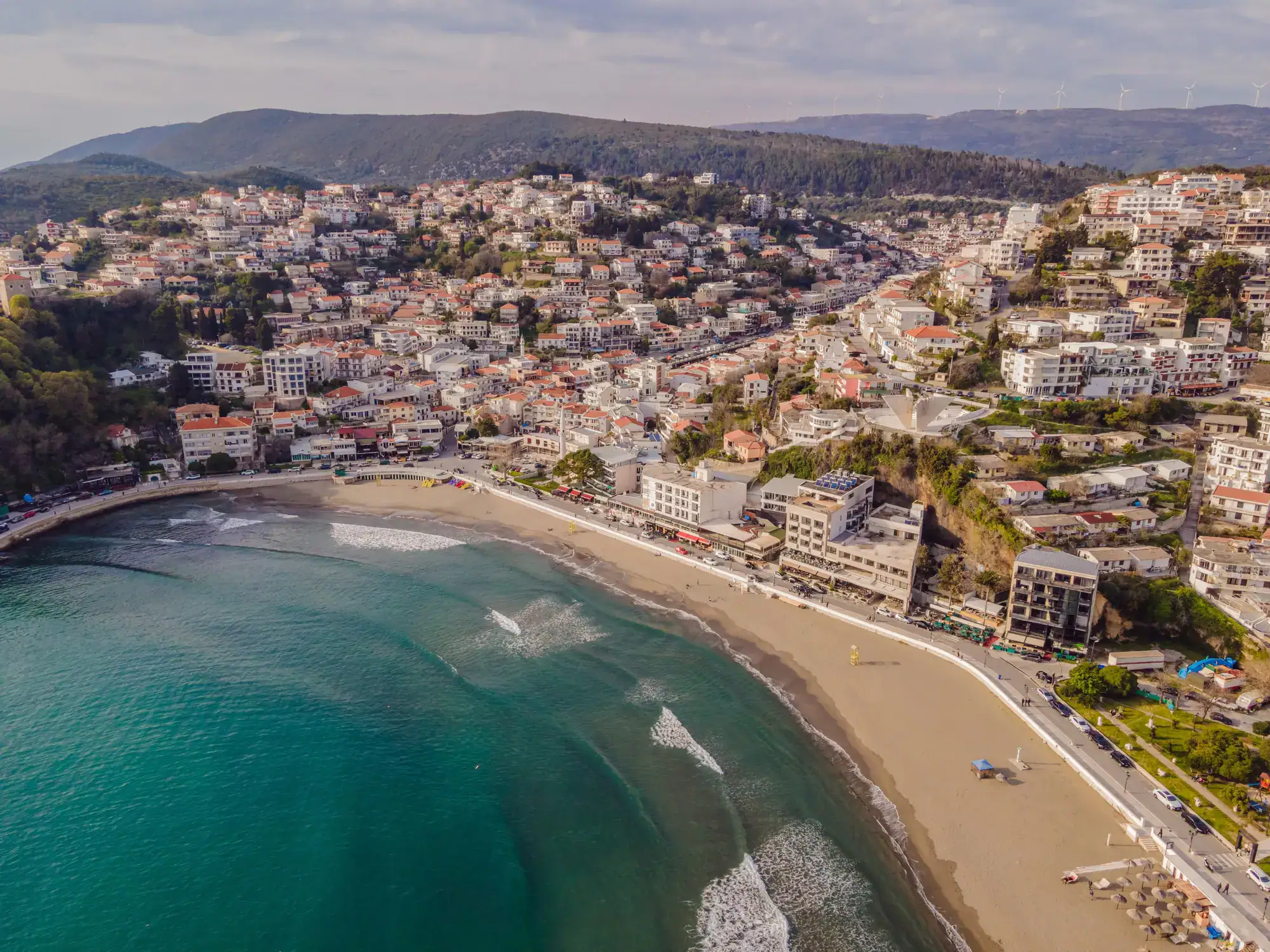 Aerial view of a light colored, curved beach at the bottom of Ulcinj with white apartment buildings and teal ocean, one of the cheapest beach destinations in Europe.