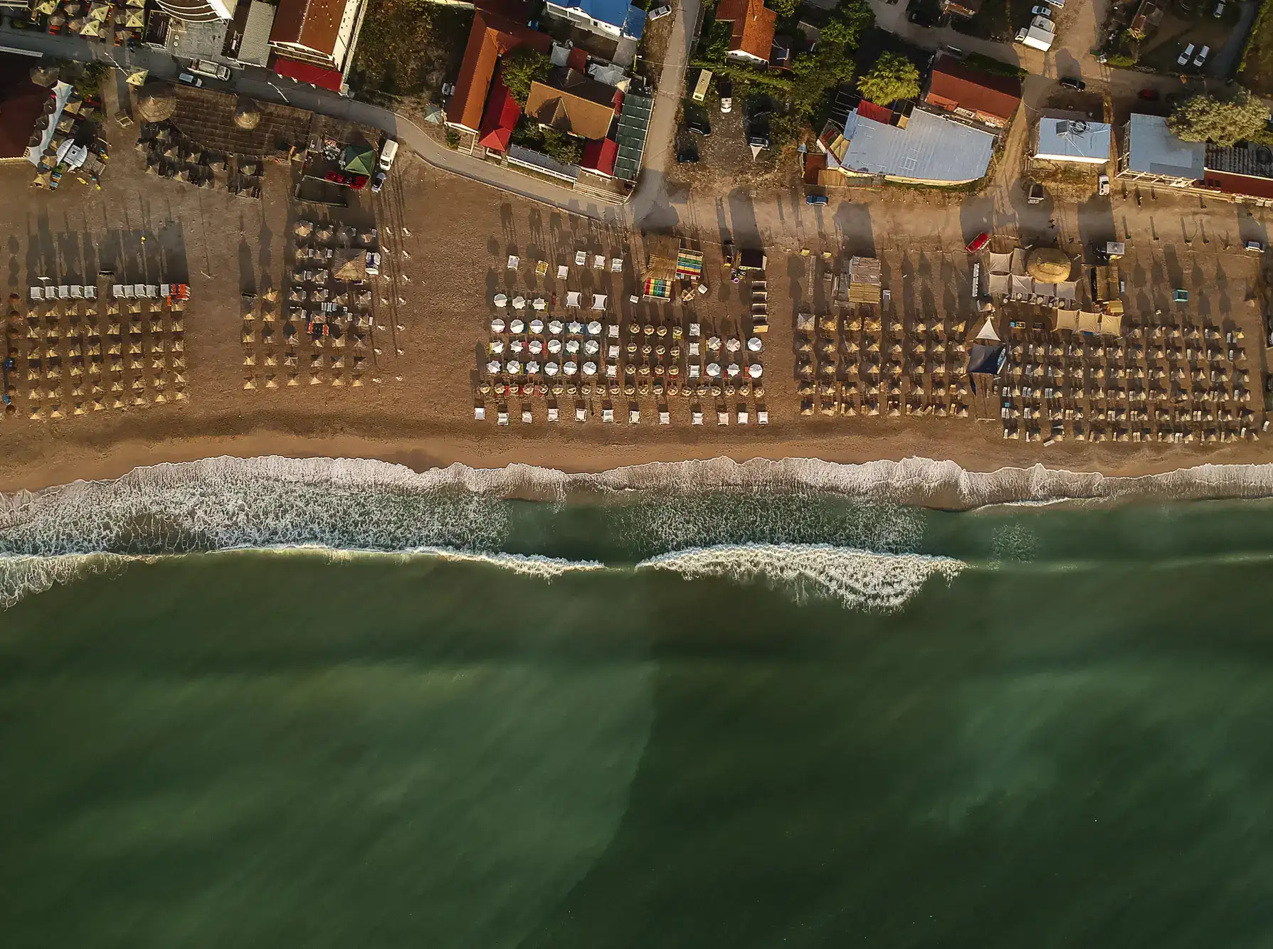 Drone view straight down on Vama Veche Beach, a wide stretch of golden sand with sections of sunbeds and parasols, green water with waves crashing in and some houses and greenery in the background. One of the most affordable beach destinations in Europe. 