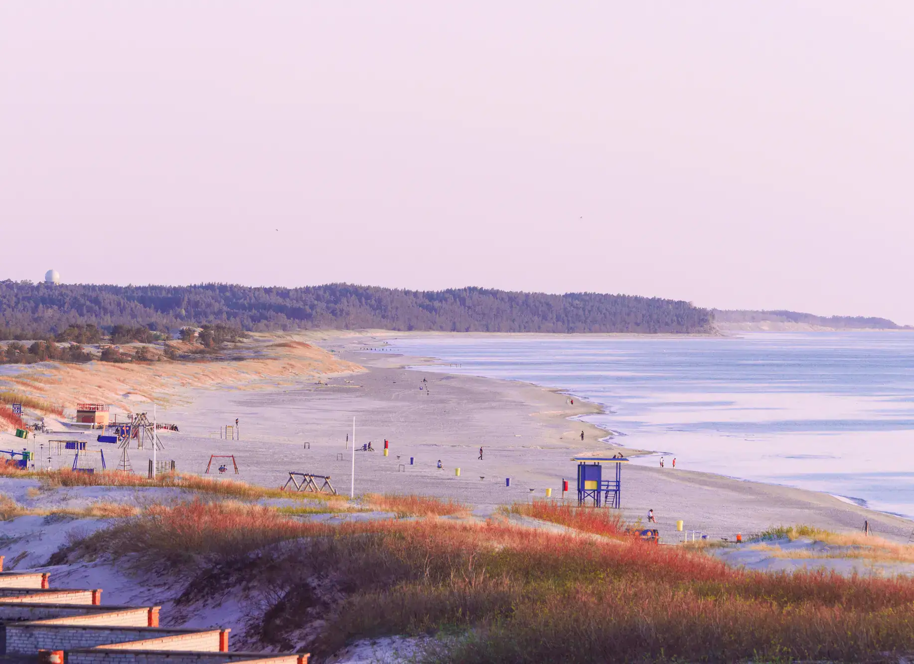 Early morning with pink light over Ventspils Beach, with sandiness covered in grass in the foreground, a blue lifeguard tower, forest in the background, one of the cheapest beach towns in Europe.