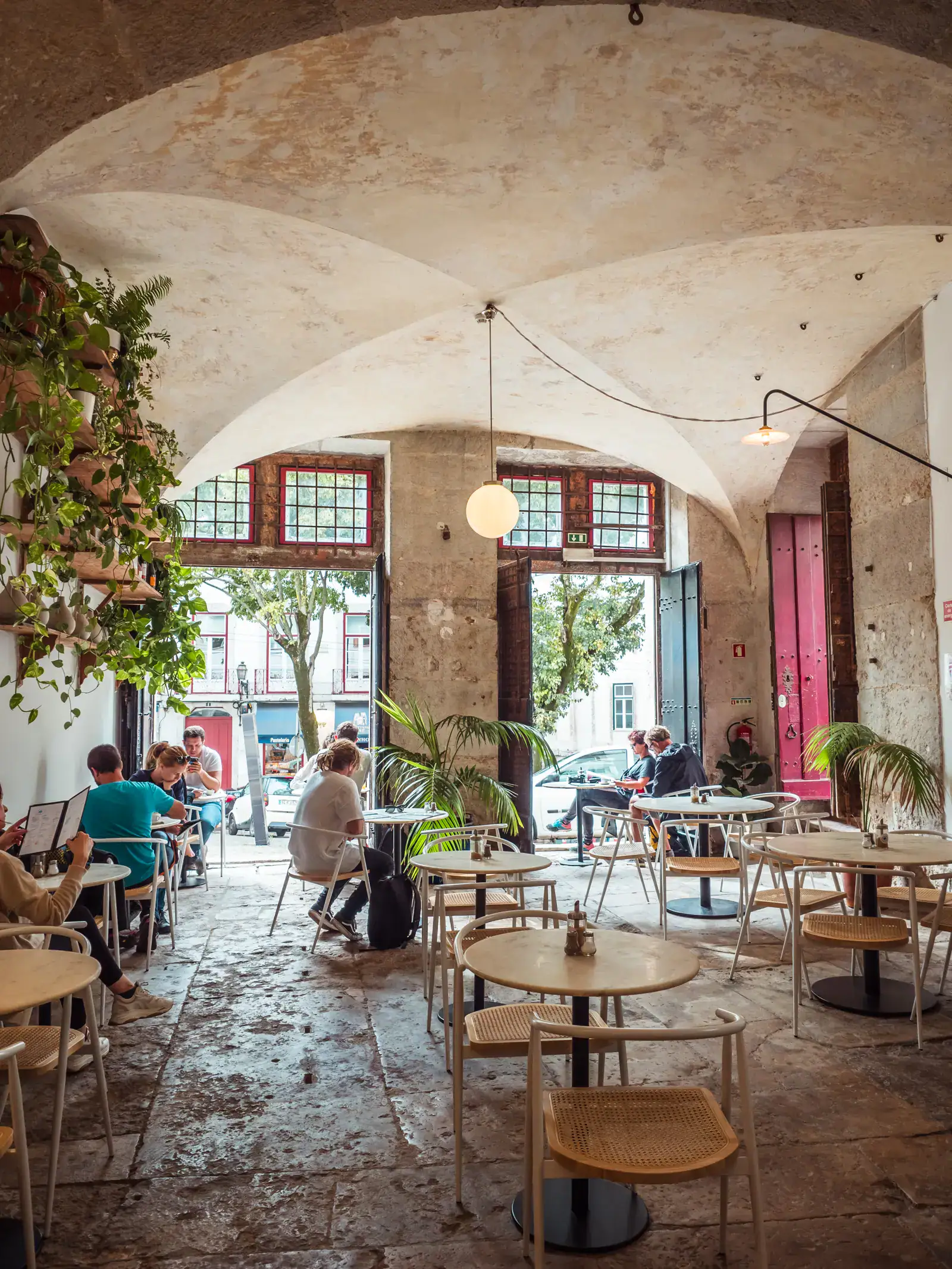 Light wooden tables and chairs inside Dear Breakfast Alfama, an old stone building with an arched roof, people sitting by the open doors, one of the best breakfast spots in Lisbon.