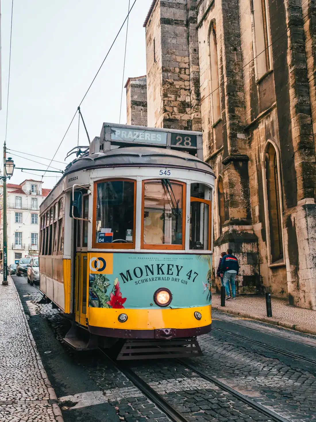The iconic yellow 28 tram running past Lisbon Cathedral, a beige stone building on the right in the photo, on a gloomy day in Lisbon.