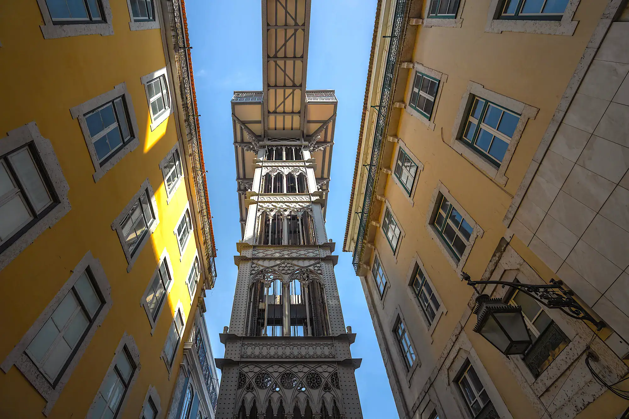 Looking up at the iconic Santa Justa Lift in Lisbon, with one old, yellow apartment building on each side on a sunny day with blue skies.