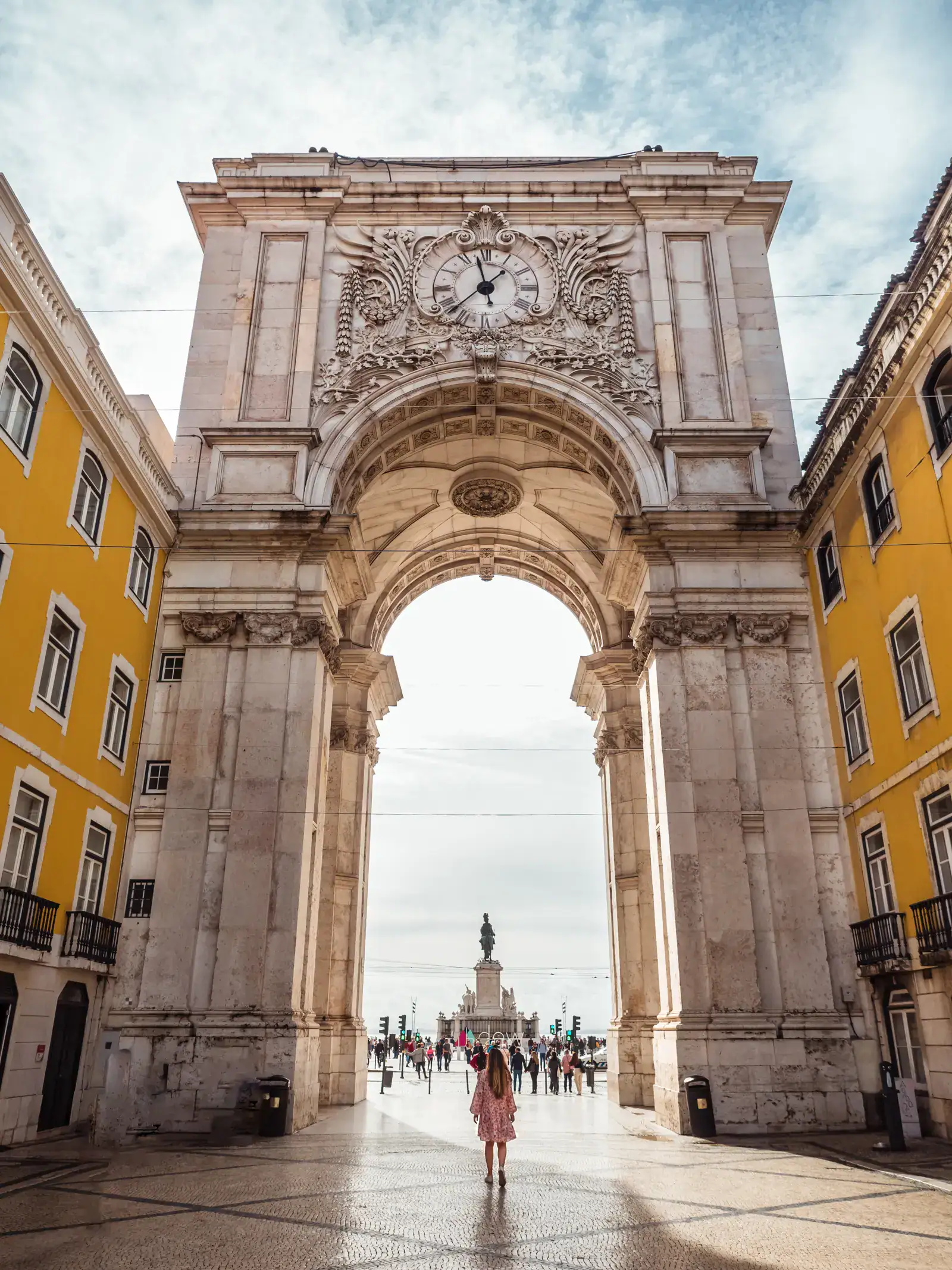 Woman with long hair, wearing a pink dress, walking alone under the huge and ornate stone Arch of Rua Augusta, with tall, yellow buildings on both sides during one day in Lisbon.