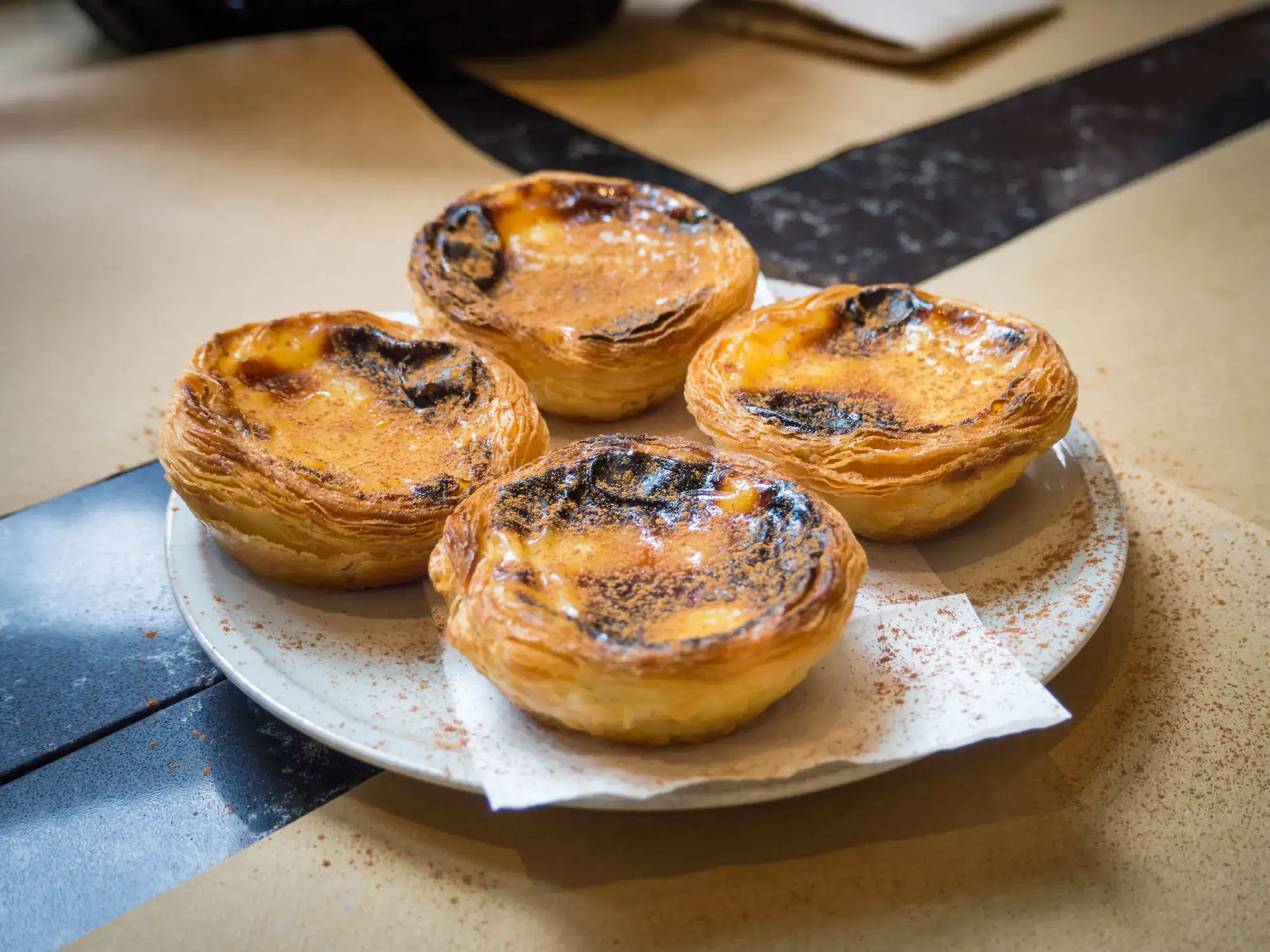 White round plate with a white napkin and four Past&eacute;is de Nata topped with cinnamon during a one-day itinerary in Lisbon, Portugal.