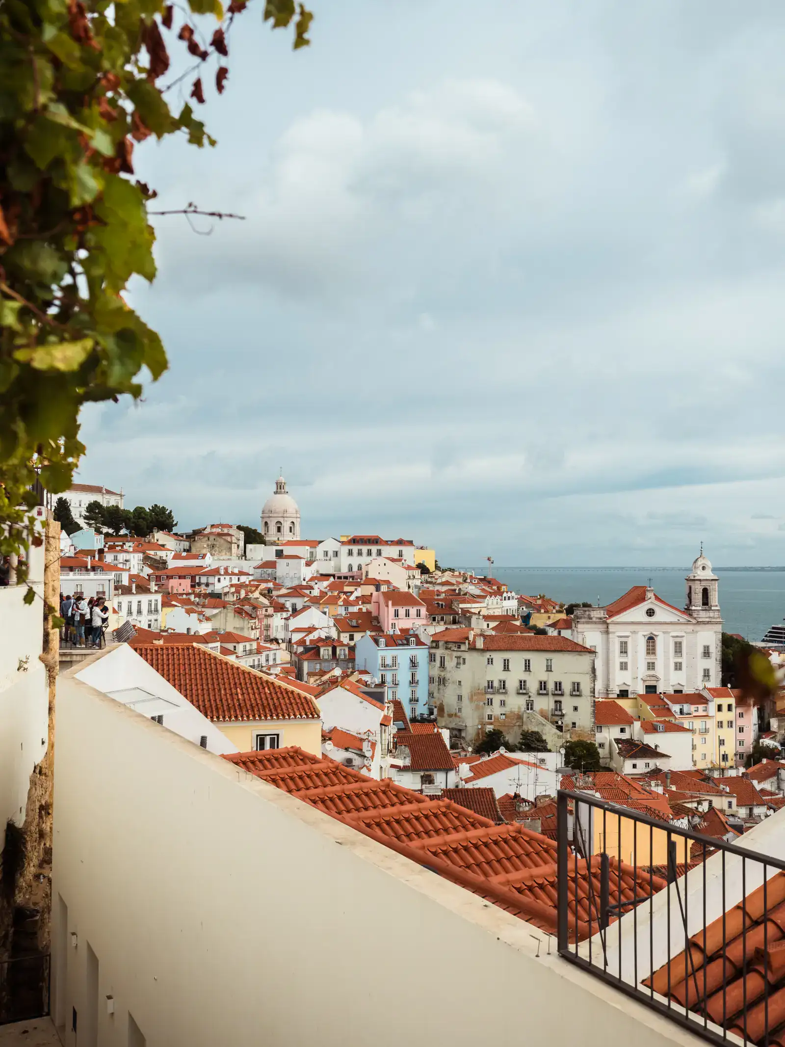 View across orange colored rooftops, pastel colored buildings and churches by the ocean from Miradouro de Santa Luzia in Lisbon, one of the top things to do in Lisbon during a one-day itinerary.