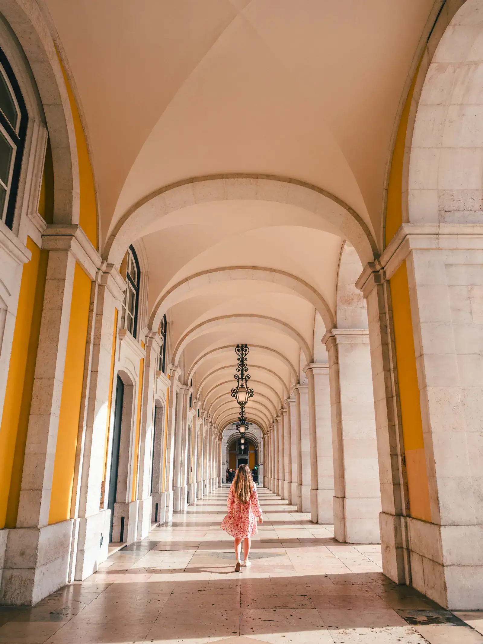 Woman with long dark blonde hair, wearing a pink dress and white shoes, walking from the camera under an arched walkway with yellow walls and white roof outside the Supreme Court of Justice, one of the highlights in Lisbon during a one day itinerary.