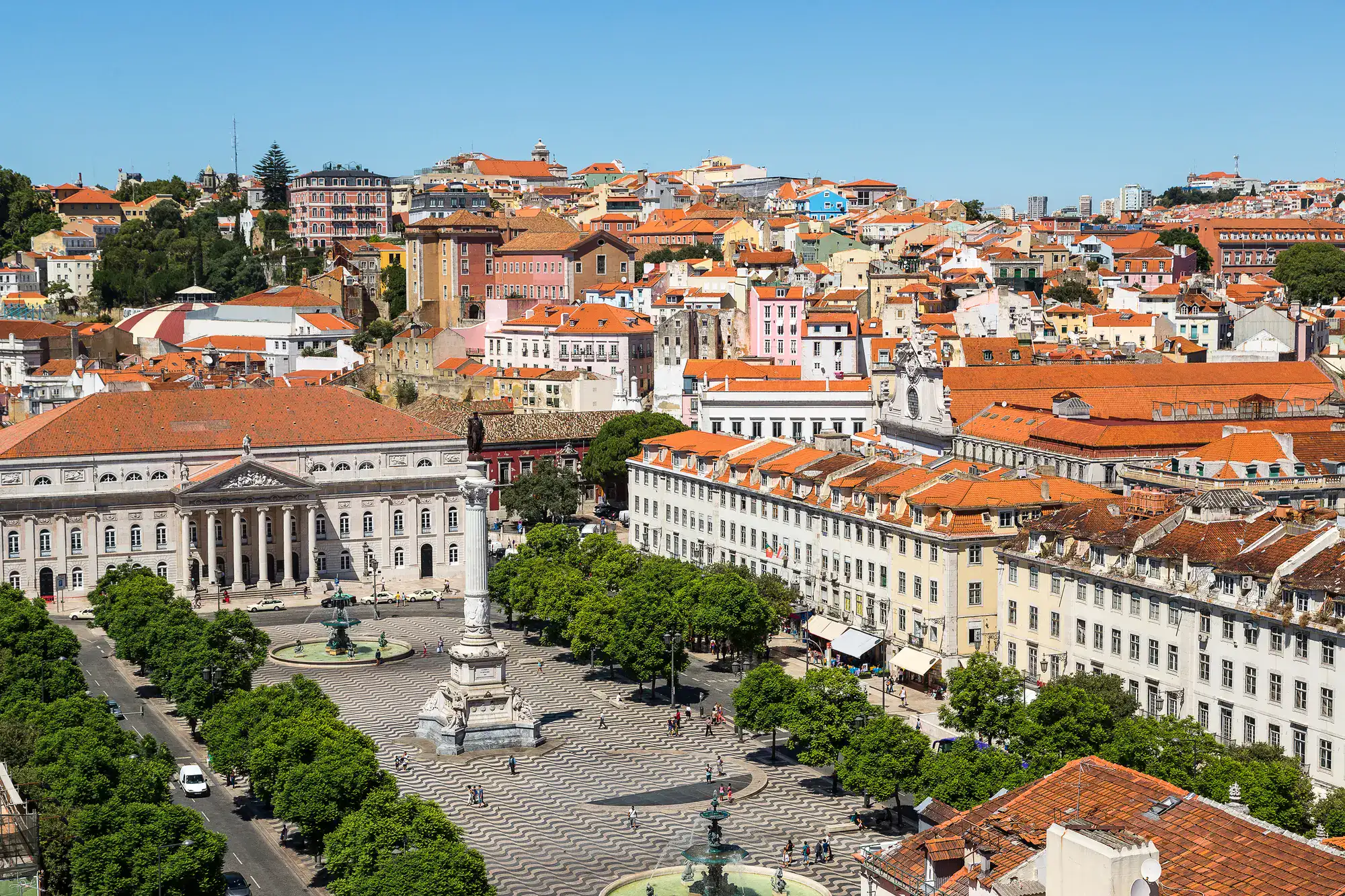 Large, tiled Rossio Square with a wave pattern in light and dark grey. Large pillar in the middle, grand building at the end with six pillars at the entrance, colorful houses with orange roofs in the background on a sunny day in Lisbon.