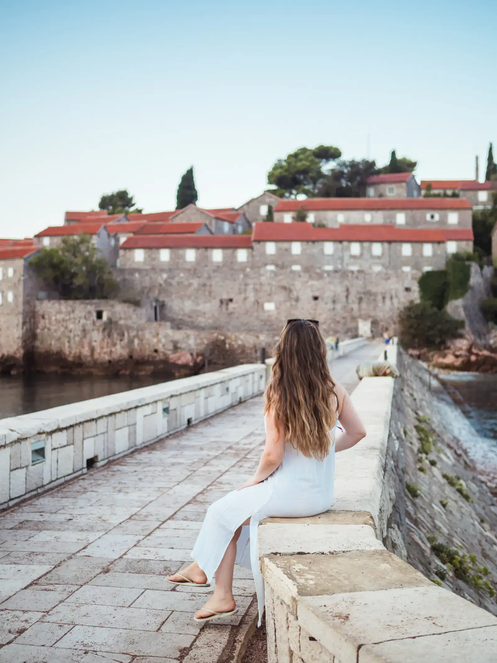 Woman with long dark blond hair, wearing a white maxi dress, sitting on the stone wall along the pathway leading to the old Sveti Stefan Island with beige stone buildings with orange roofs, early in the morning in Sveti Stefan Montenegro.