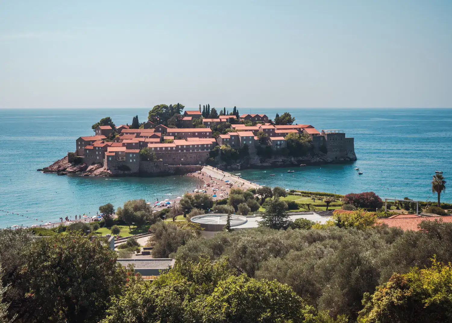 Looking from a viewpoint in the circular Sveti Stefan Island with old stone houses with orange roofs and some green trees surrounded by turquoise water, stone path leading to it from Sveti Stefan Beach with a green park closest to the camera on a sunny day in Sveti Stefan Montenegro. 