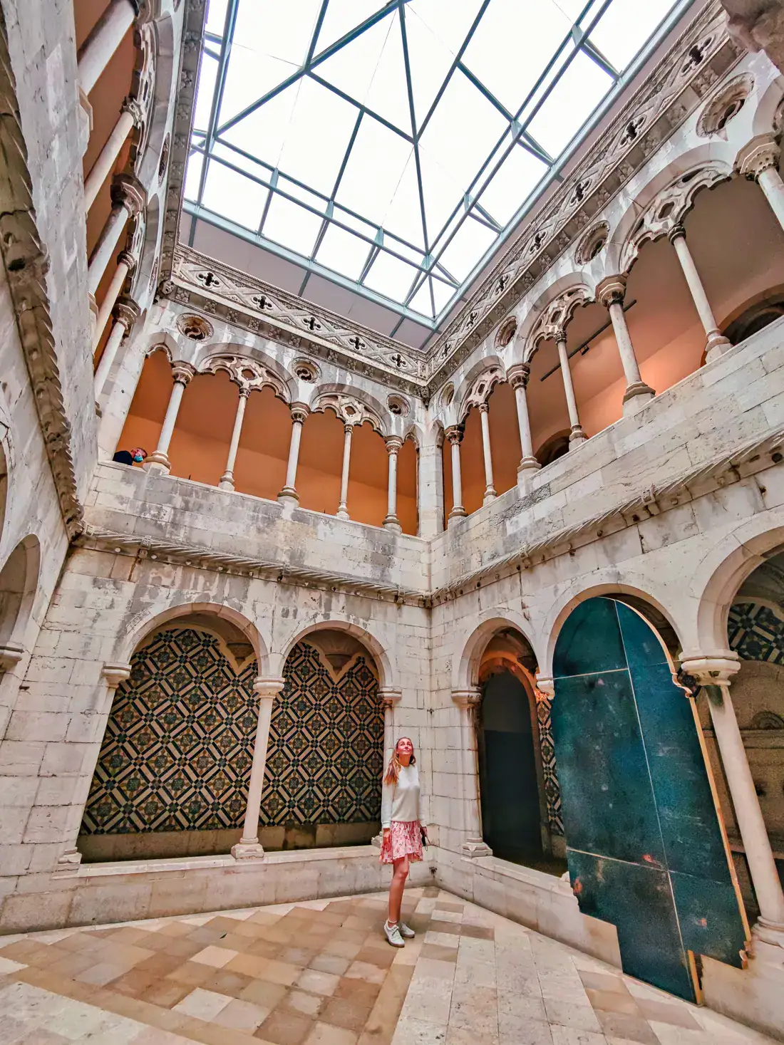 Woman wearing a pink skirt, white sweater and white shoes standing inside a grand room with arches, columns and a glass pyramid ceiling in Azulejo Tile Museum in Lisbon during a first-timers itinerary.