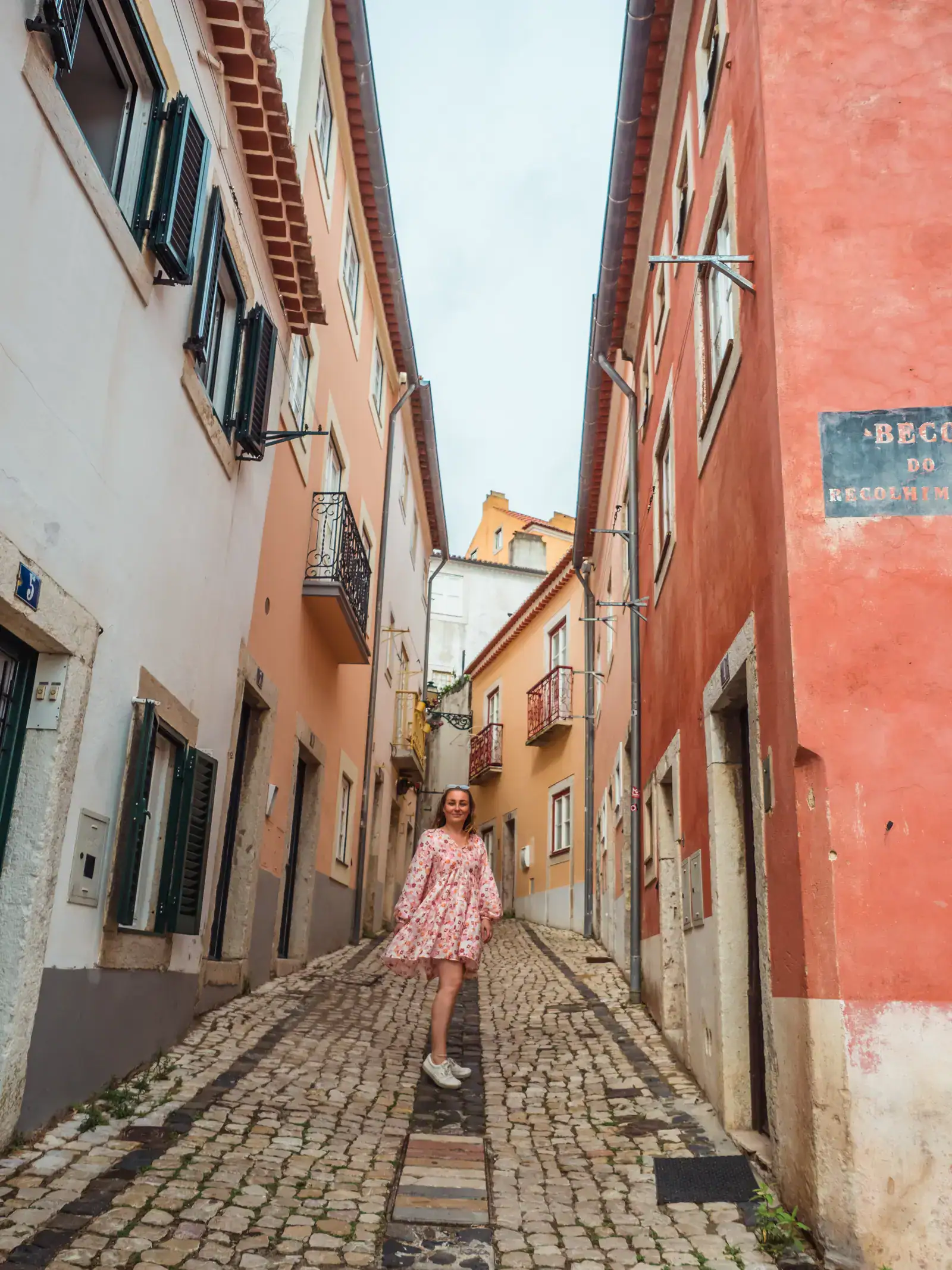 Woman wearing a long sleeve, flowy pink patterned dress, walking up a cobbled hill in Lisbon, between red, white and orange old stone buildings during the perfect one day Lisbon itinerary.