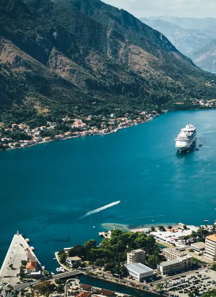 View from high up on the Old Town walls in Kotor of a cruise ship coming in on teal blue water between tall mountains. Is Kotor still worth visiting?