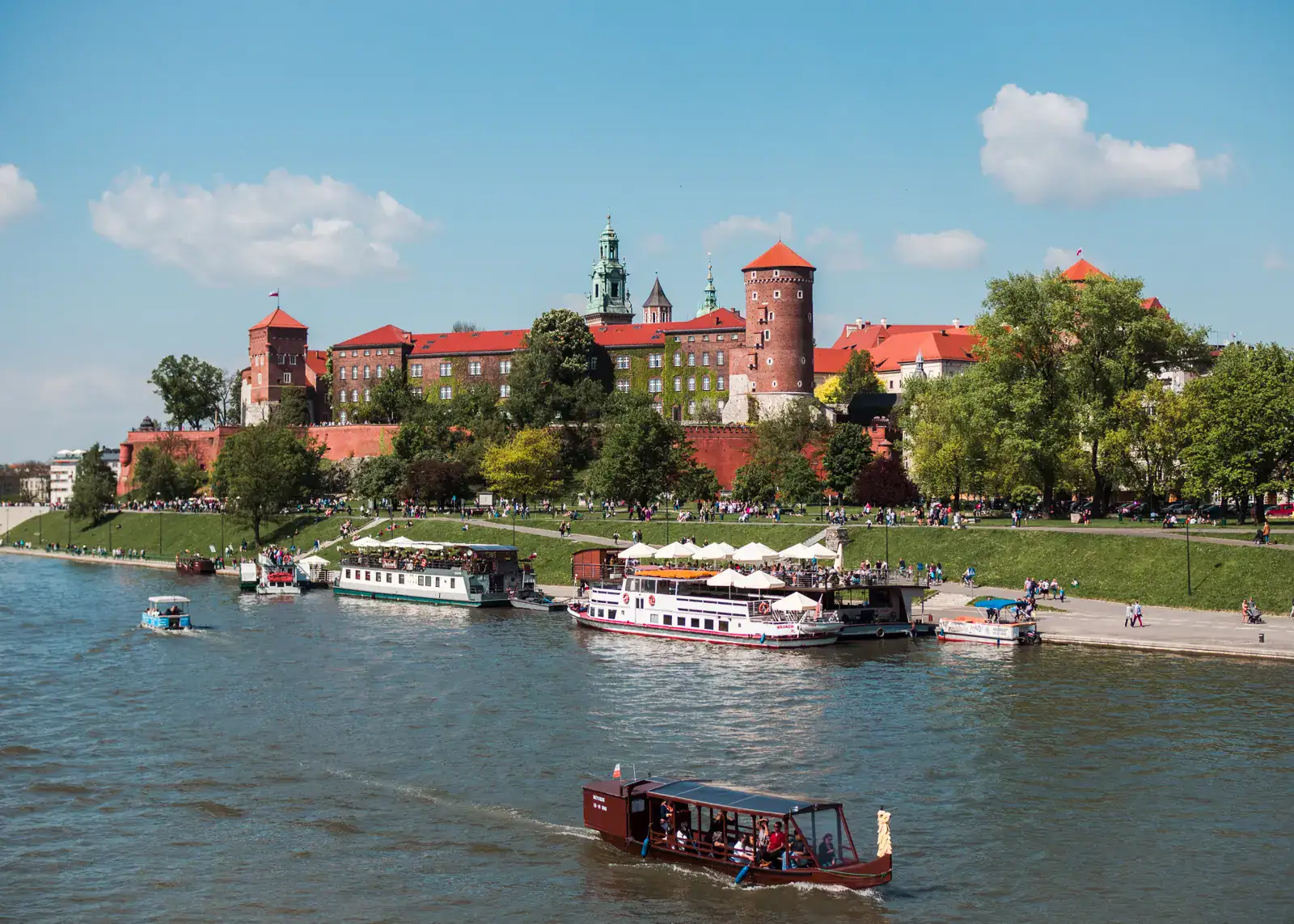 View of the Vistula River with a brown wooden boat in the foreground, two white passenger boats docked along the bank, the majestic Wawel Castle in the background with orange roof and two towers, surrounded by greenery, on a sunny day. Best day trips from Krakow.