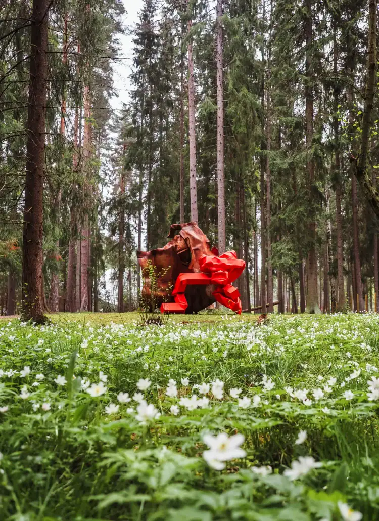 View of a red and brown contemporary metal sculpture in the forest, seen from a the ground in a meadow of white flowers and greenery at Kistefos Museum during a day trip from Oslo.