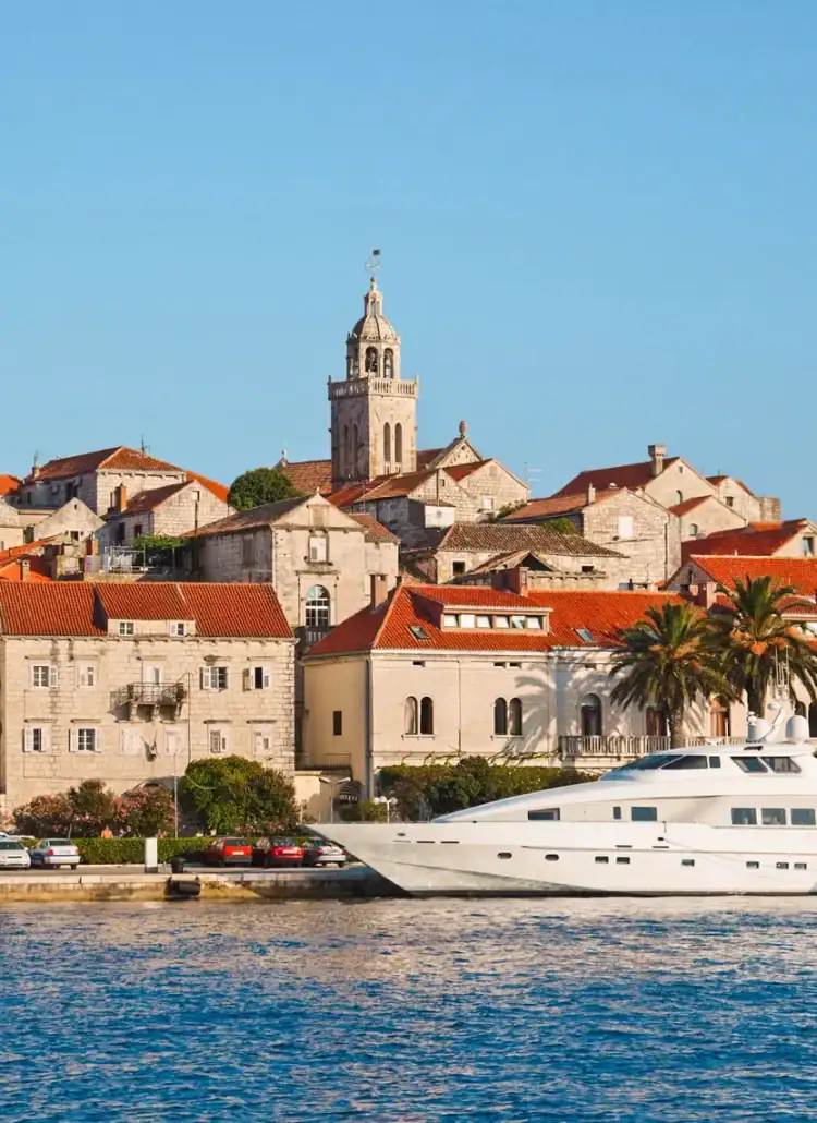 White yacht docked at the edge of Korcula Town with a beige stone tower, beige stone houses with orange roofs and Aminess Younique Korcula Heritage Hotel with palm trees in front on a sunny day with blue skies. Where to stay on Korcula.