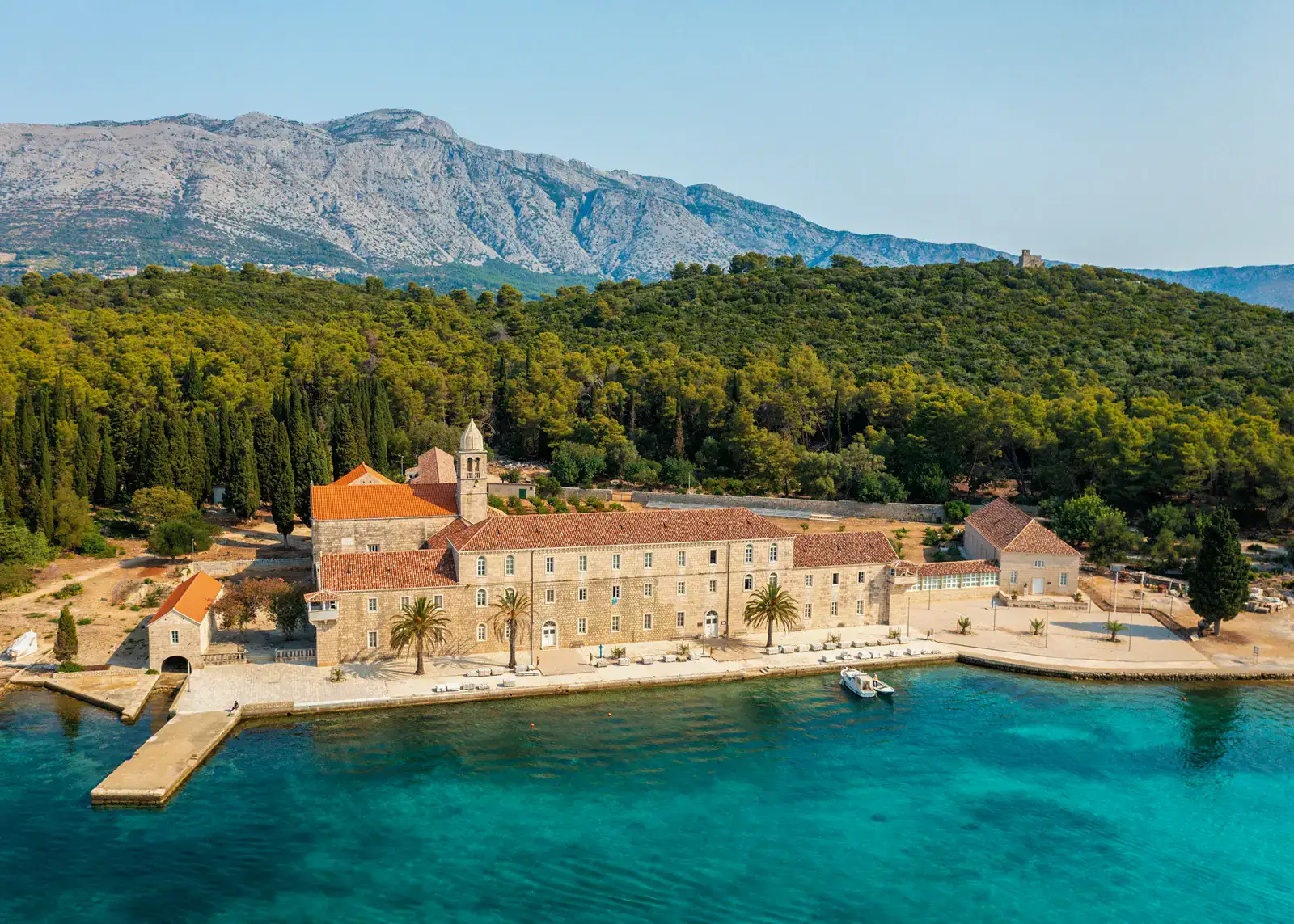Drone view of a large monastery in beige sandstone with orange roof, three palm tress in front, green forest and a mountain in the background. deep turquoise water in the foreground, seen on Badija Island, during island hopping in Korcula.