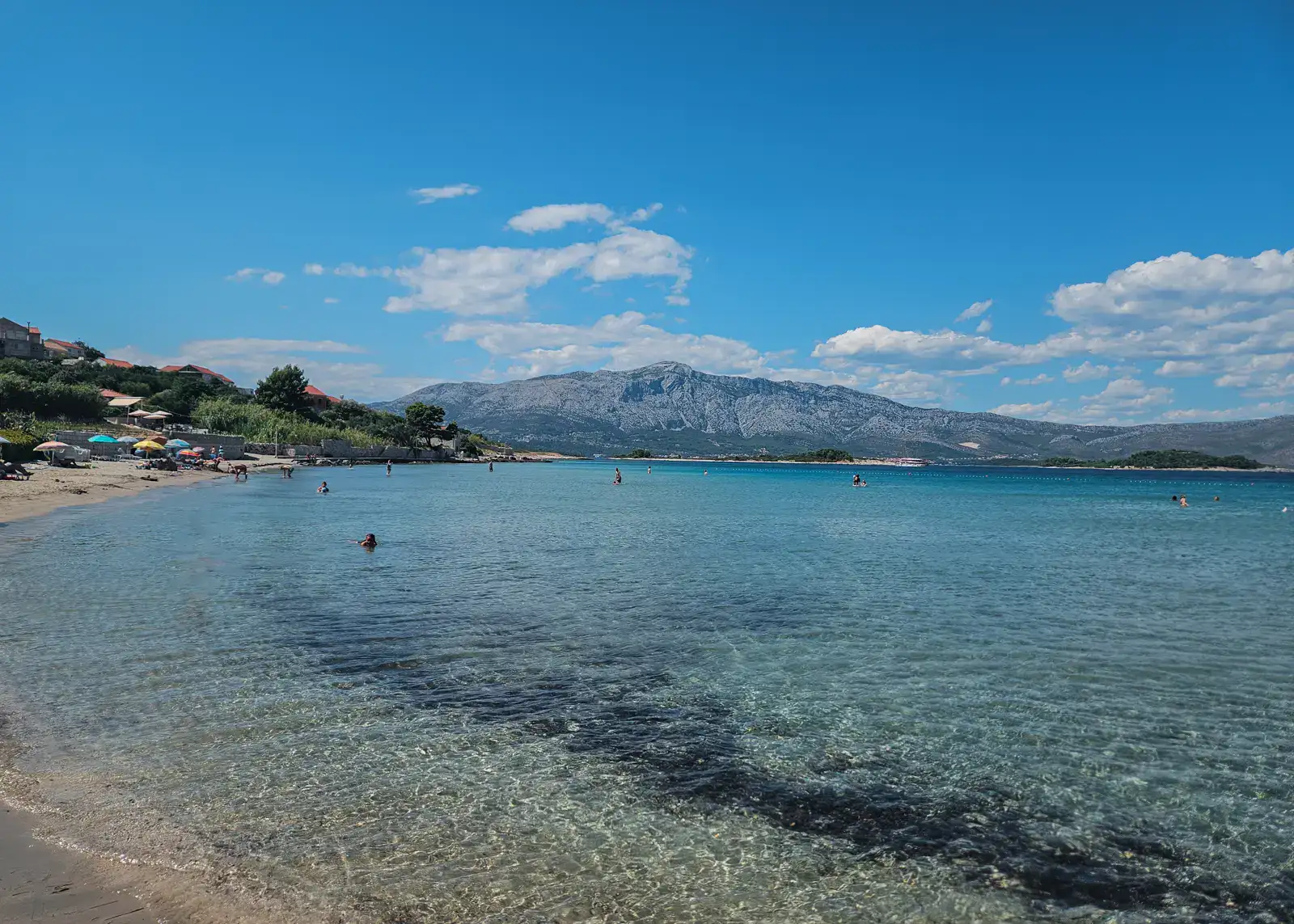 View of Bilin Zal Beach, a shallow bay of turquoise water, a narrow strip of sand with colorful parasols on the left and a mountain in the background, one of the best beaches on Korcula.