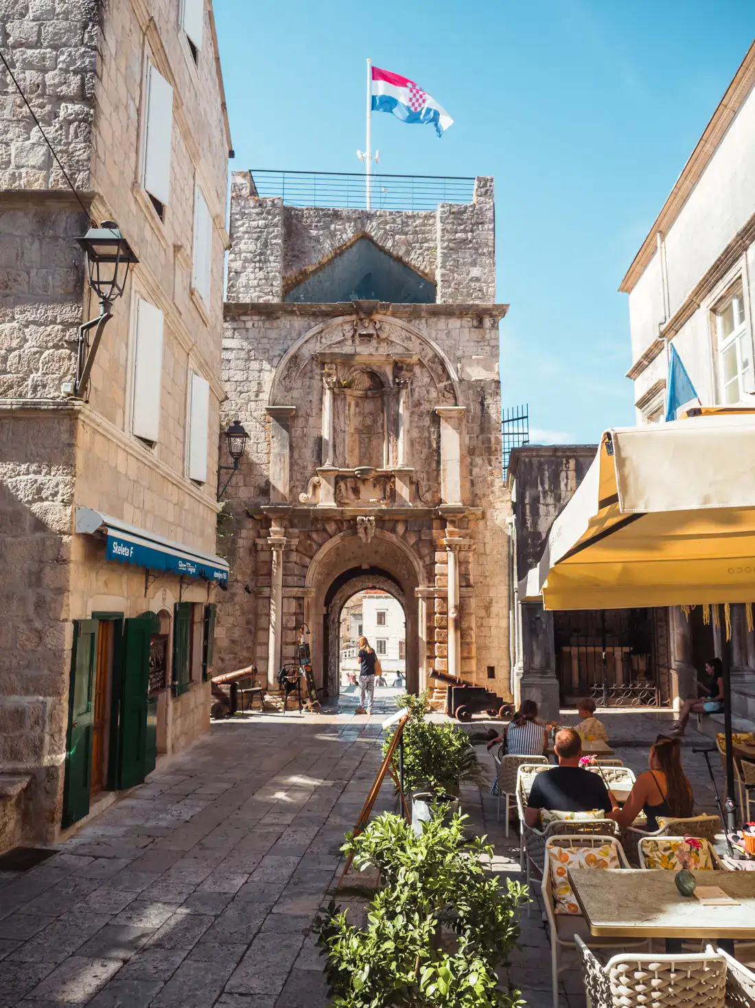 Looking towards the tower entrance of Korcula Old Town, restaurant under an yellow awning on the right, store in the stone building on the left on a sunny day.