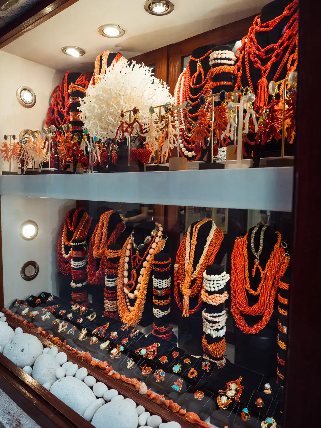 Luxurious window display of a jewelry store selling coral jewelry, many necklaces in red, orange and coral tones on black displays, large white coral in the middle of the top shelf, rings and pendants in the bottom, shopping in Korcula Town.